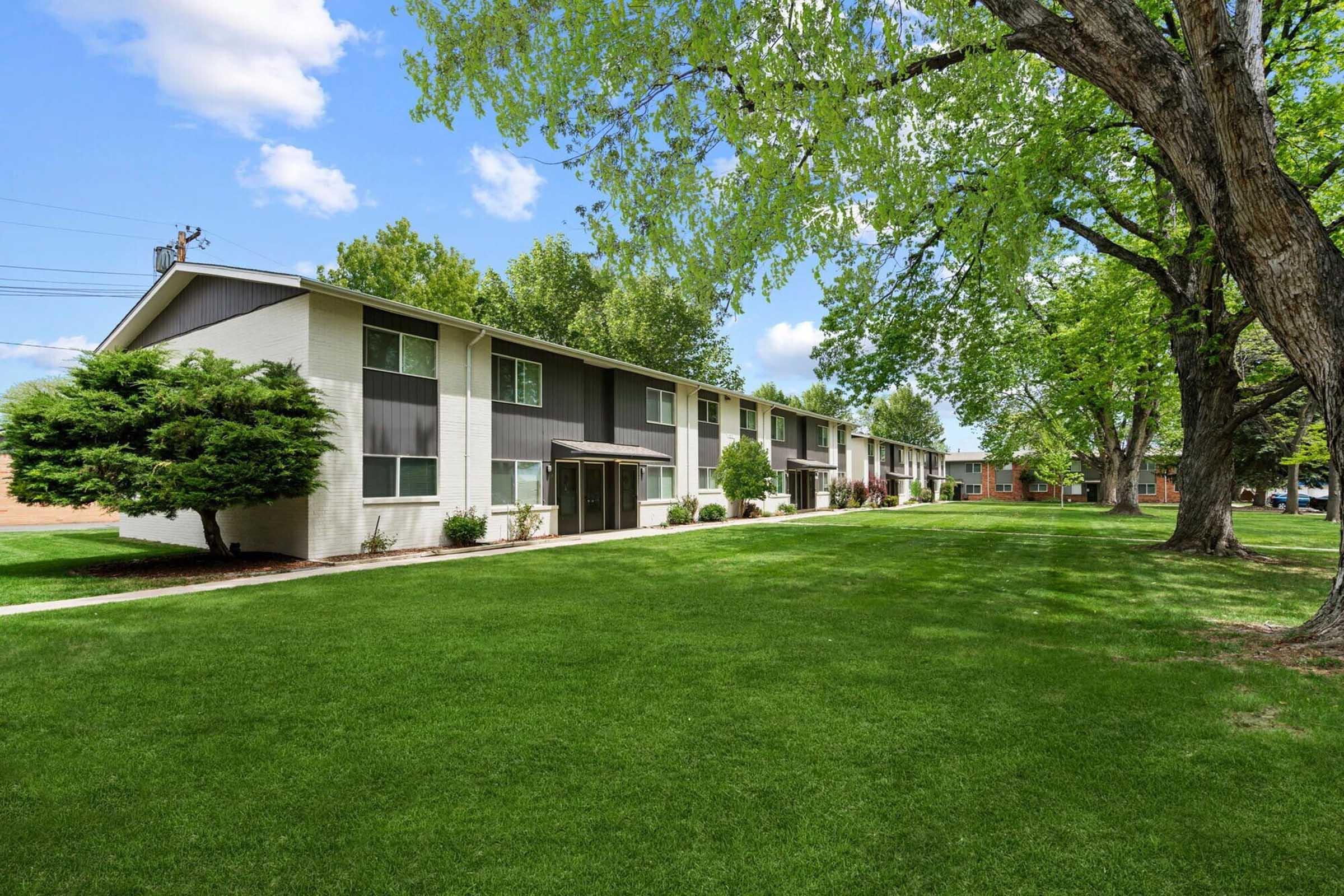 A grassy area with a row of multi-story residential buildings surrounded by trees. The buildings have a mix of white and dark exteriors. The scene is bright with blue skies and fluffy white clouds, creating a serene and well-maintained atmosphere.
