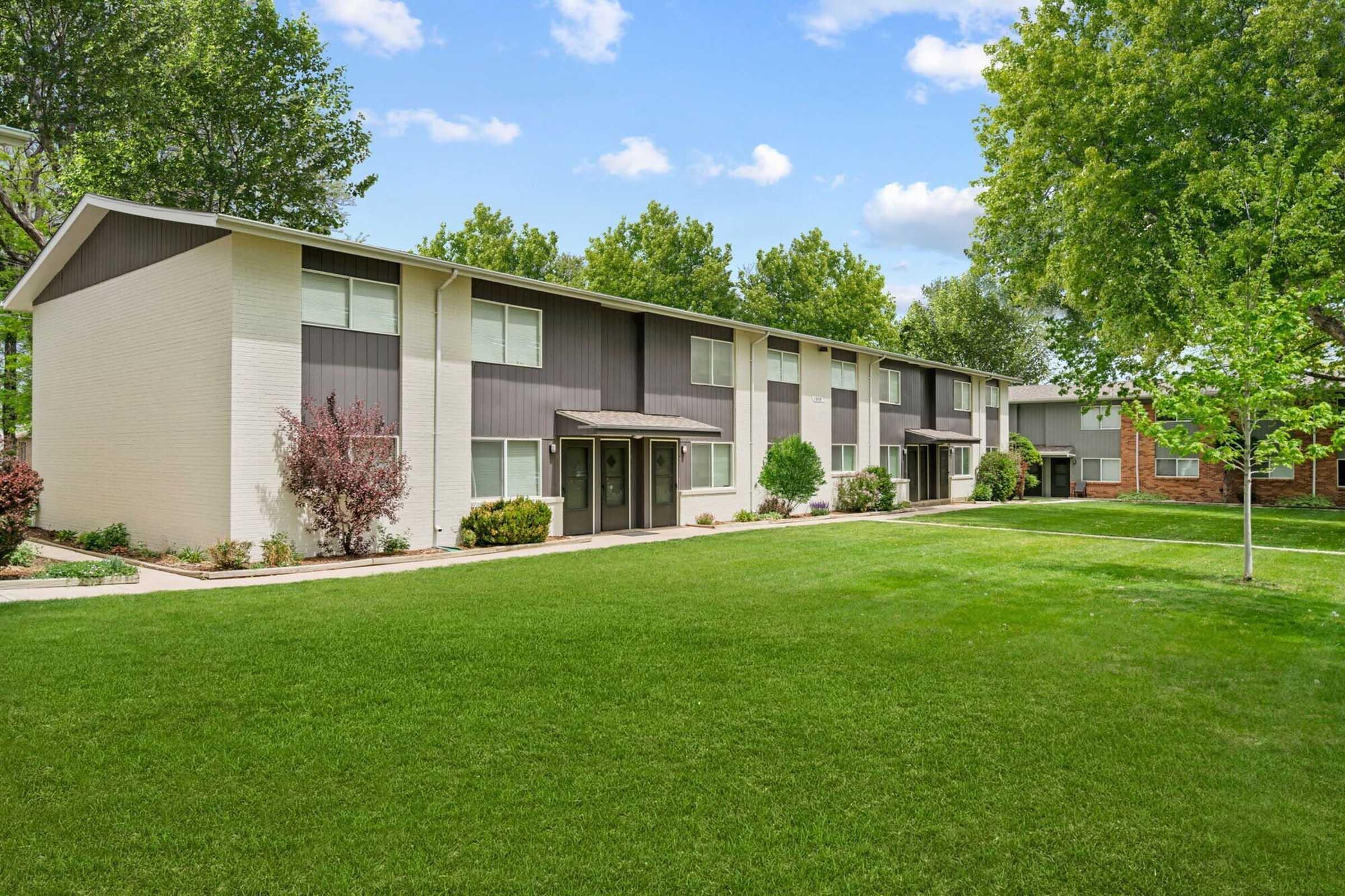 A modern apartment complex featuring two-story buildings with a blend of light-colored and dark gray exteriors. The well-maintained landscape includes a lush green lawn with trees and shrubs, creating a welcoming outdoor environment. Clear blue sky and fluffy clouds are visible in the background.