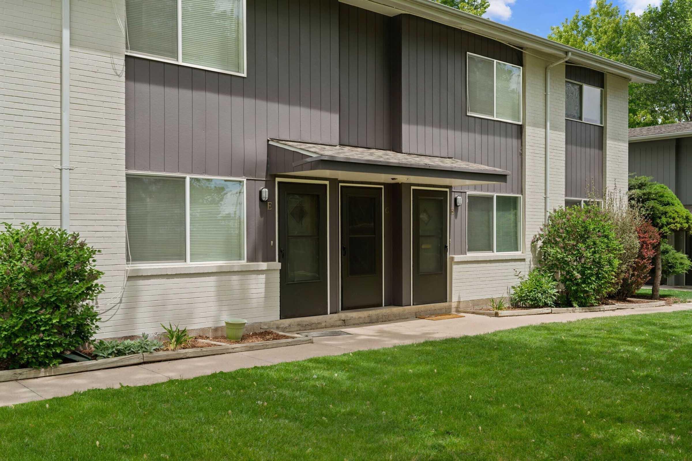 View of a two-story apartment building featuring a combination of gray and white siding. Two doors are located centrally, with a concrete pathway leading up to them. Surrounding the entrance are small shrubs and a well-maintained lawn with green grass. Trees are visible in the background.