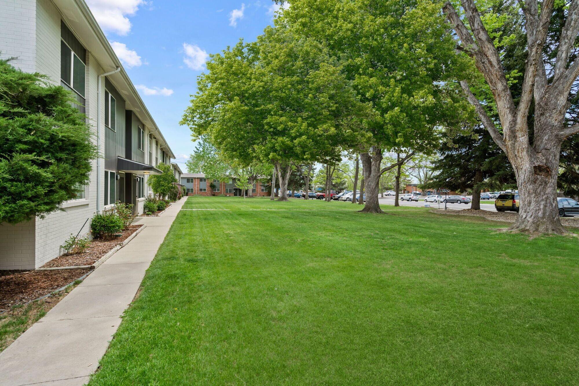 A well-maintained grassy lawn lined with trees and residential buildings. Pathways lead through the landscape, with cars parked in the background under a clear blue sky. The scene is tranquil and inviting, showcasing a pleasant outdoor space.