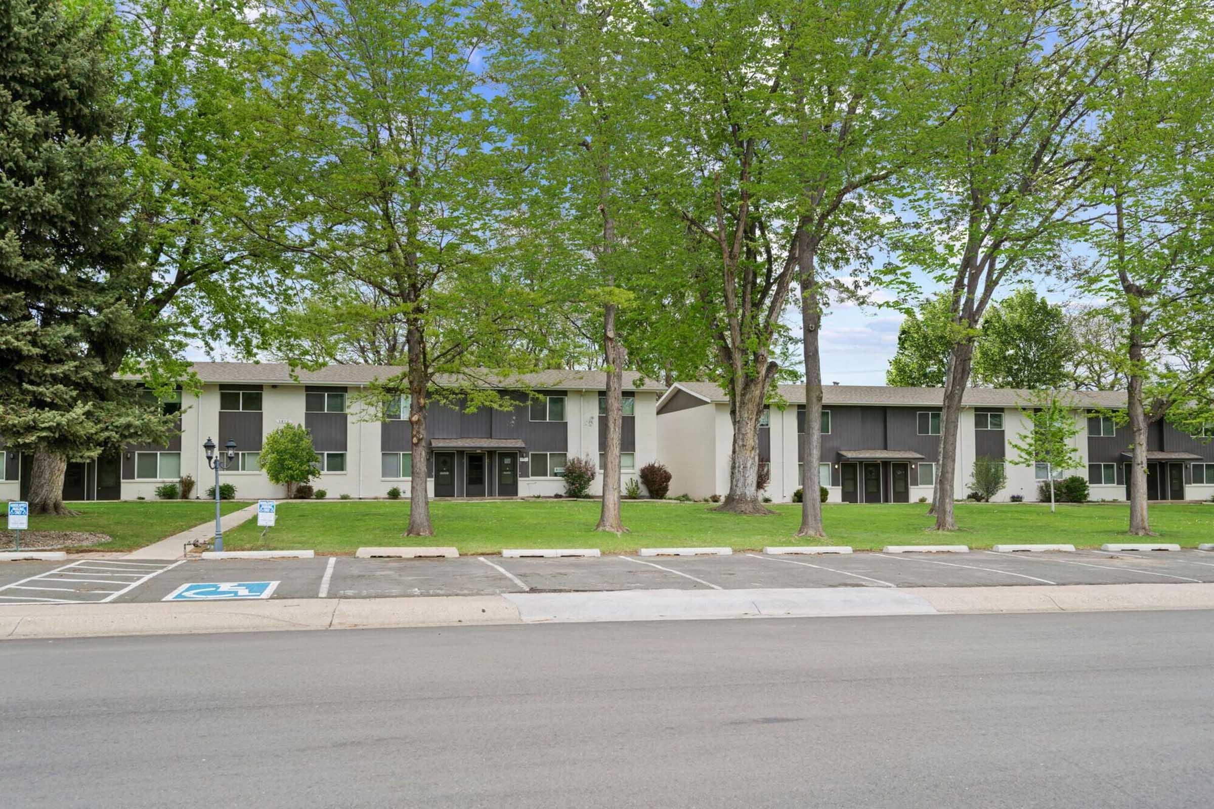 A modern residential building with multiple units, featuring large windows and gray exterior panels. The property is surrounded by lush green trees and a well-maintained lawn. Visible parking spaces are in front, with one designated for disabled parking. The scene is set on a clear day with blue skies.