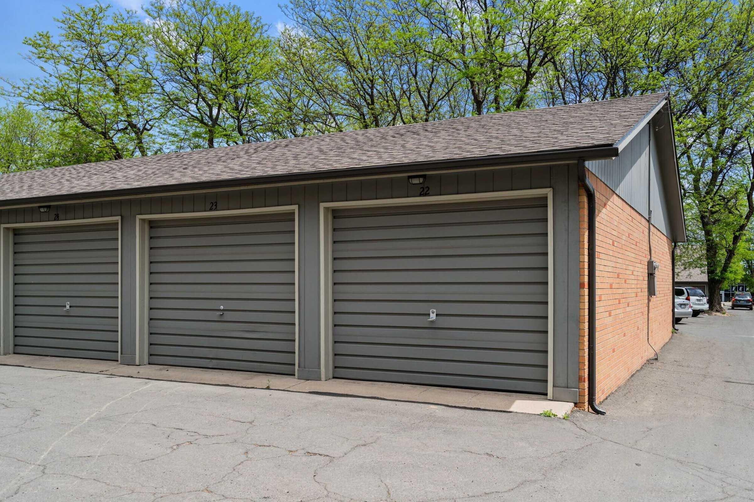 A brick building with multiple garage doors, featuring grey siding and a sloped roof. The image shows two garage doors, labeled "21" and "22," set against a backdrop of trees with green leaves and a clear blue sky. The ground is asphalt, with visible parking spaces nearby.