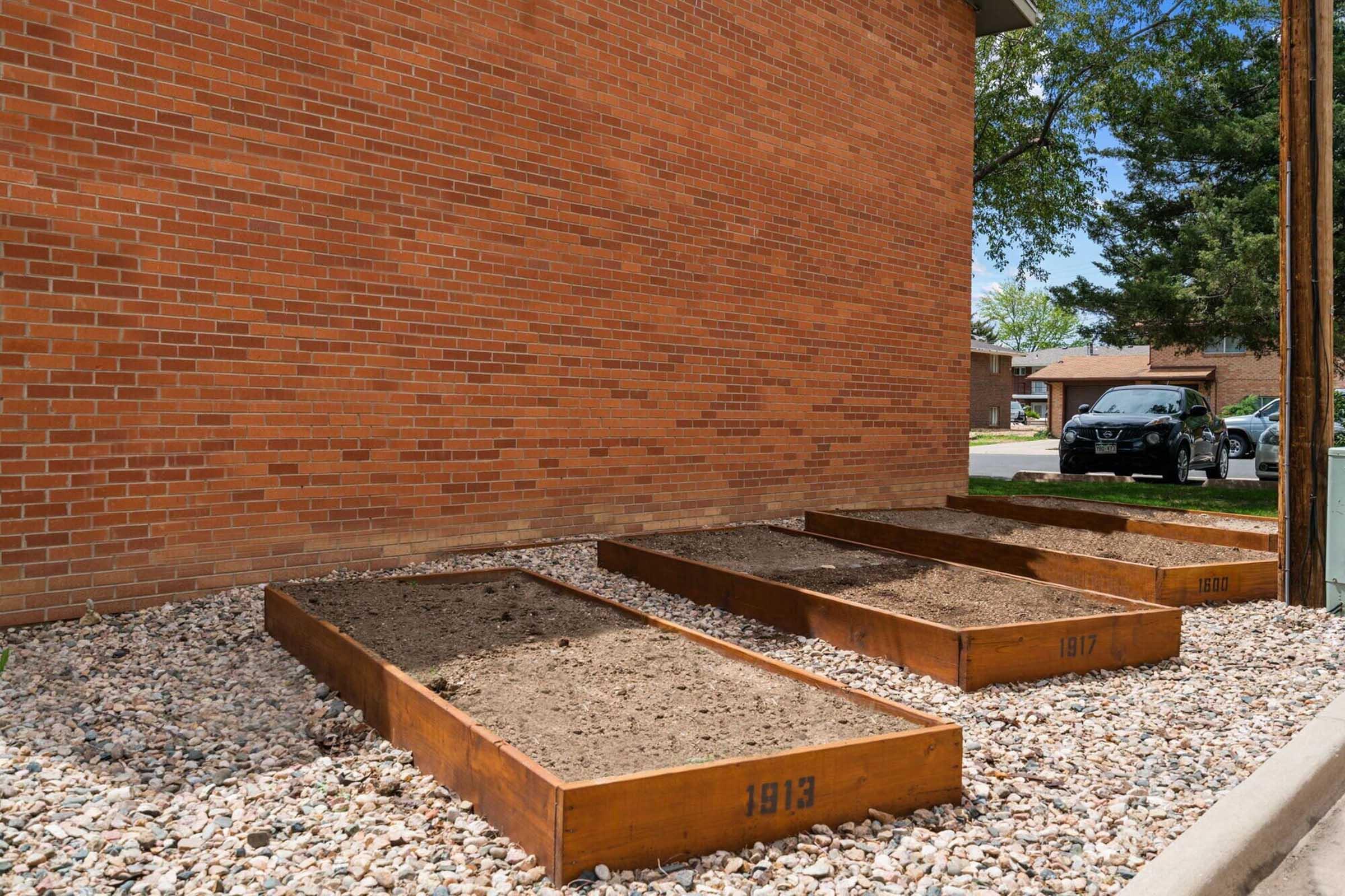 Three empty wooden planting boxes labeled "1913," "1917," and "2010," arranged on a gravel surface next to a brick wall. A black car is parked in the background, and green trees are visible in the vicinity.