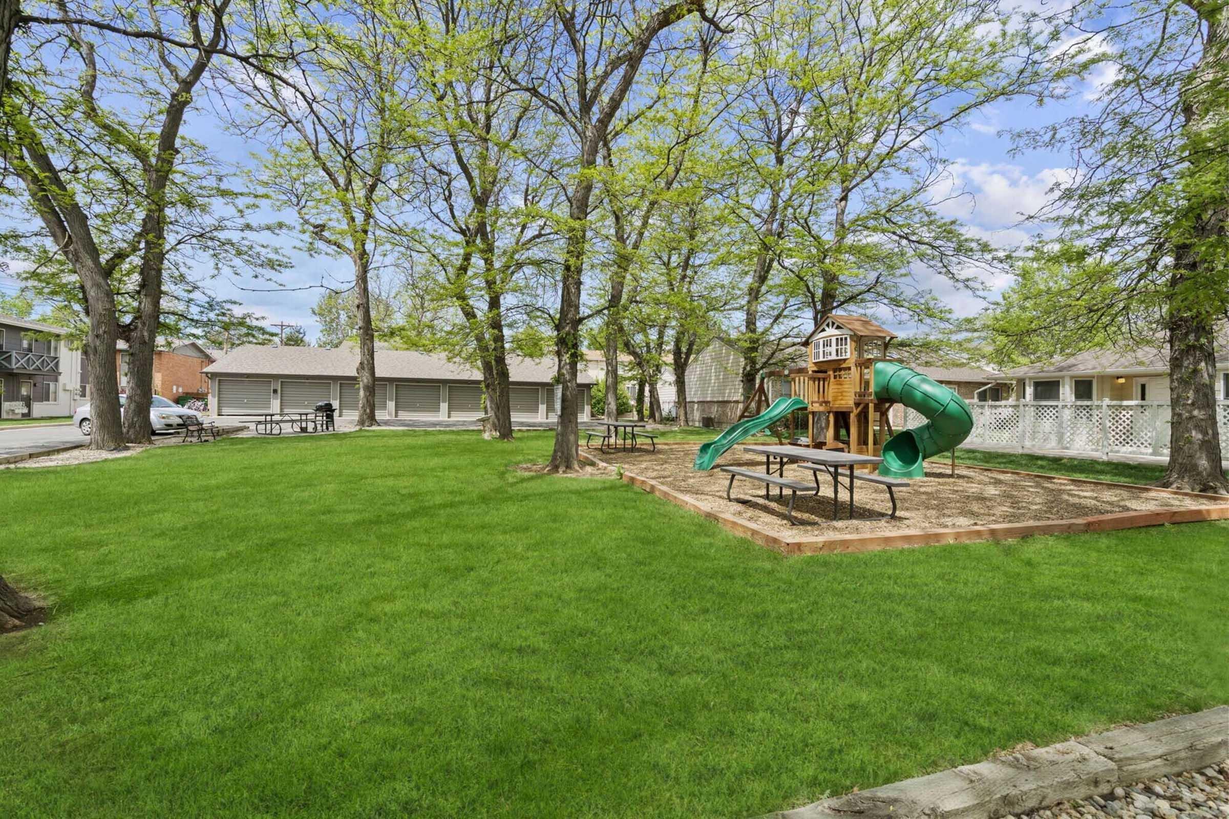 A grassy park area featuring playground equipment with a slide and a playhouse. Surrounding the playground are several trees providing shade. In the background, there are picnic tables and a parking lot with garages visible. The scene is set on a sunny day with a blue sky.