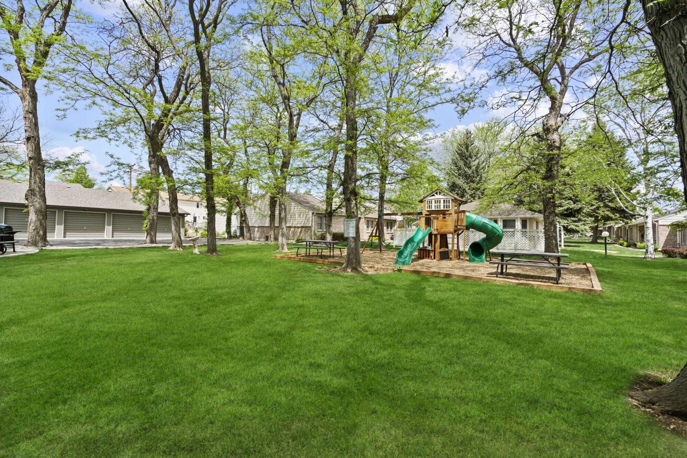 A spacious grassy area featuring a playground with a slide, surrounded by trees. Several picnic tables are placed nearby, and residential buildings are visible in the background under a clear blue sky.