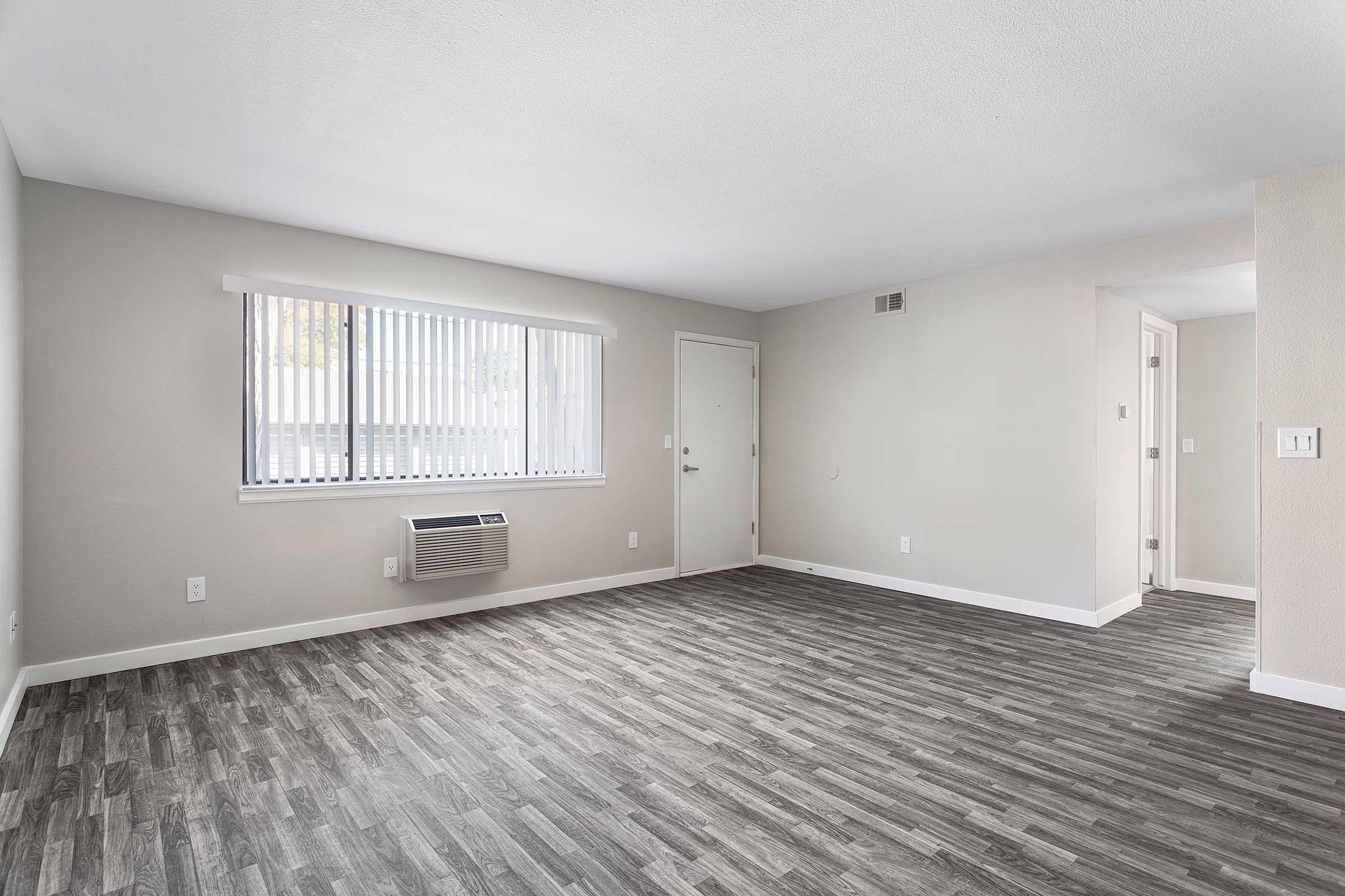 A spacious, empty room featuring a light gray wall, a window with vertical blinds, and a door. The floor is covered with wood-like laminate tiles, and there is an air conditioning unit mounted on the wall. The overall atmosphere is bright and clean, ideal for personal customization.
