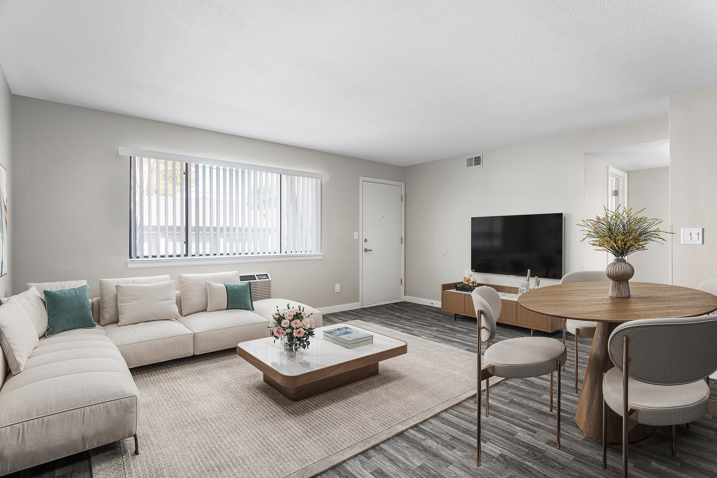 A modern living room featuring a light-colored sectional sofa, a coffee table with flowers, and a round dining table with chairs. The walls are painted in a soft gray, and there are large windows with blinds. A TV is mounted on one wall, and the floor has wood-like laminate.