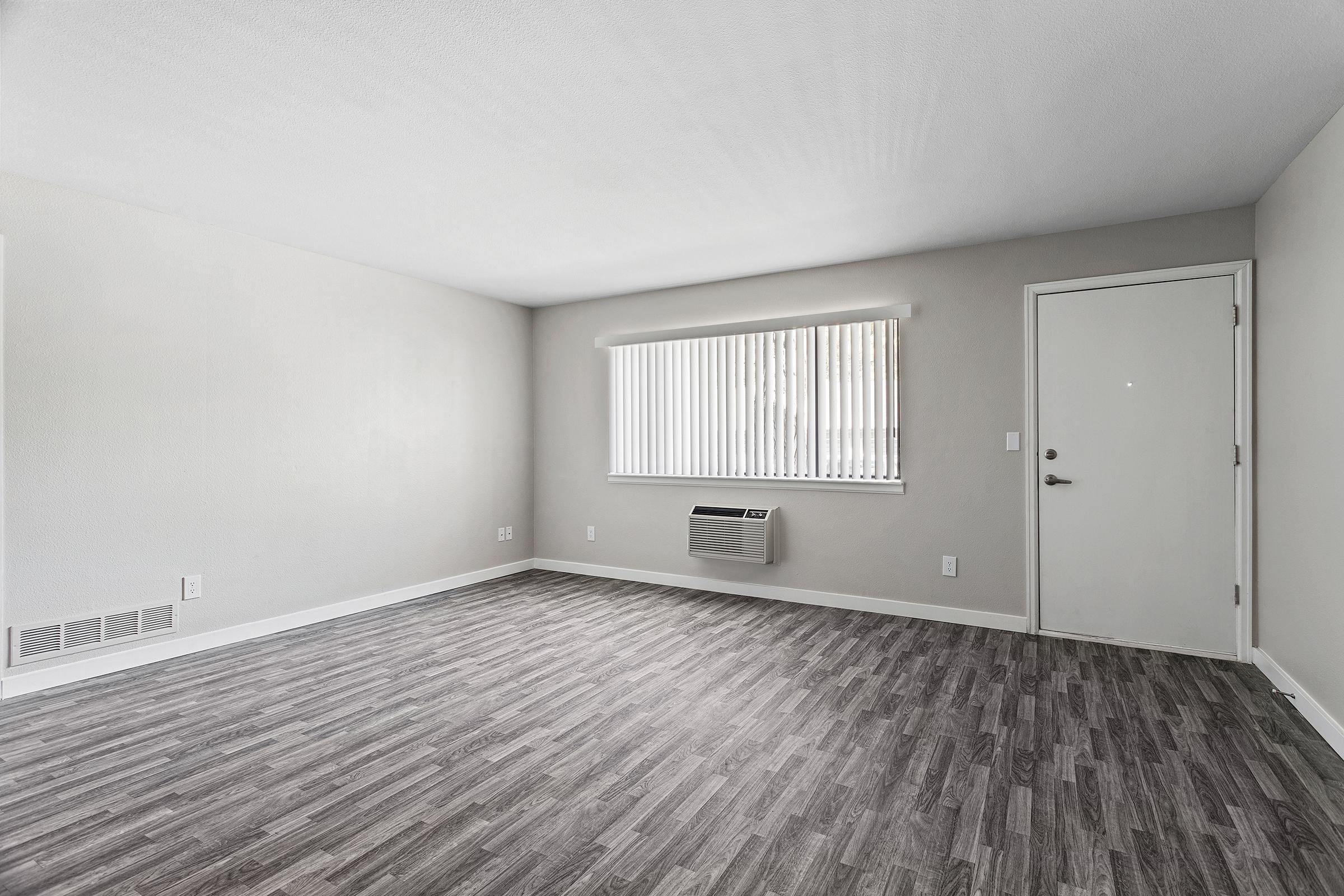 A spacious, unfurnished room with light gray walls and wood laminate flooring. A window with vertical blinds allows natural light, while an air conditioning unit is visible. The door on the right leads outside, and the overall feel is modern and minimalist.