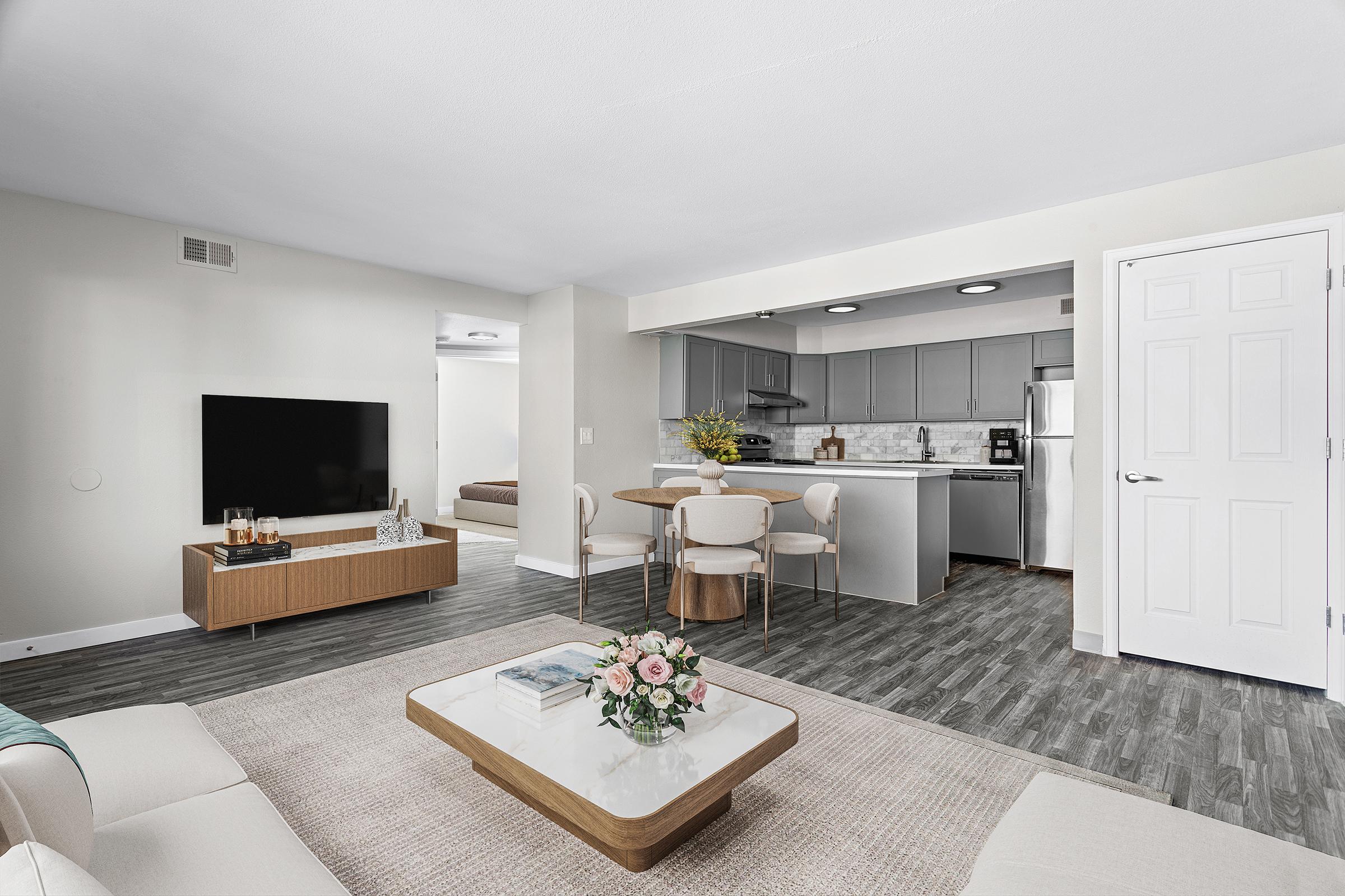 A modern, open-concept living space featuring a cozy living room with a TV on a wooden stand, a stylish coffee table with flowers, and a dining area with seating for four. The kitchen in the background has gray cabinetry and stainless steel appliances, with a light and airy overall atmosphere.