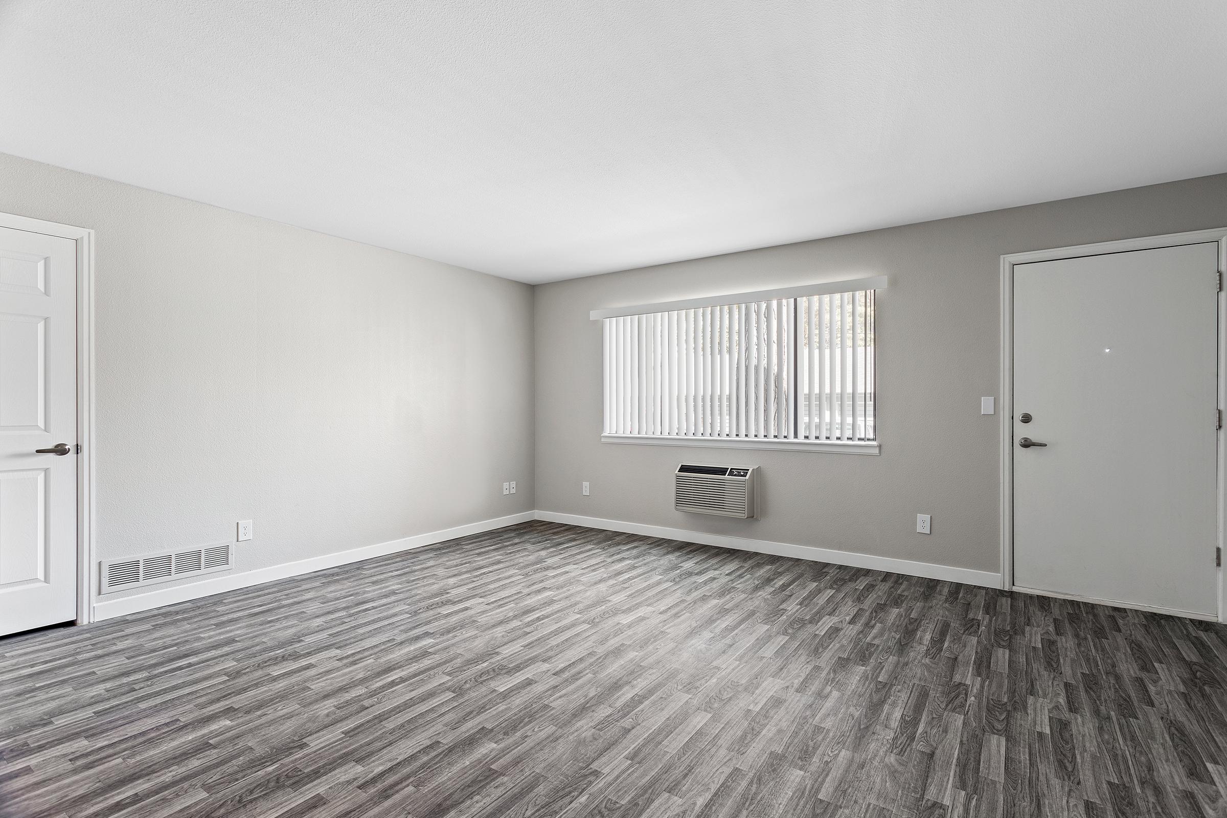 A bright, empty room with light gray walls and a window covered by vertical blinds. The floor features dark gray laminate planks. An air conditioning unit is mounted under the window, and there is a white door on the right. The overall atmosphere is clean and modern, suitable for various uses.