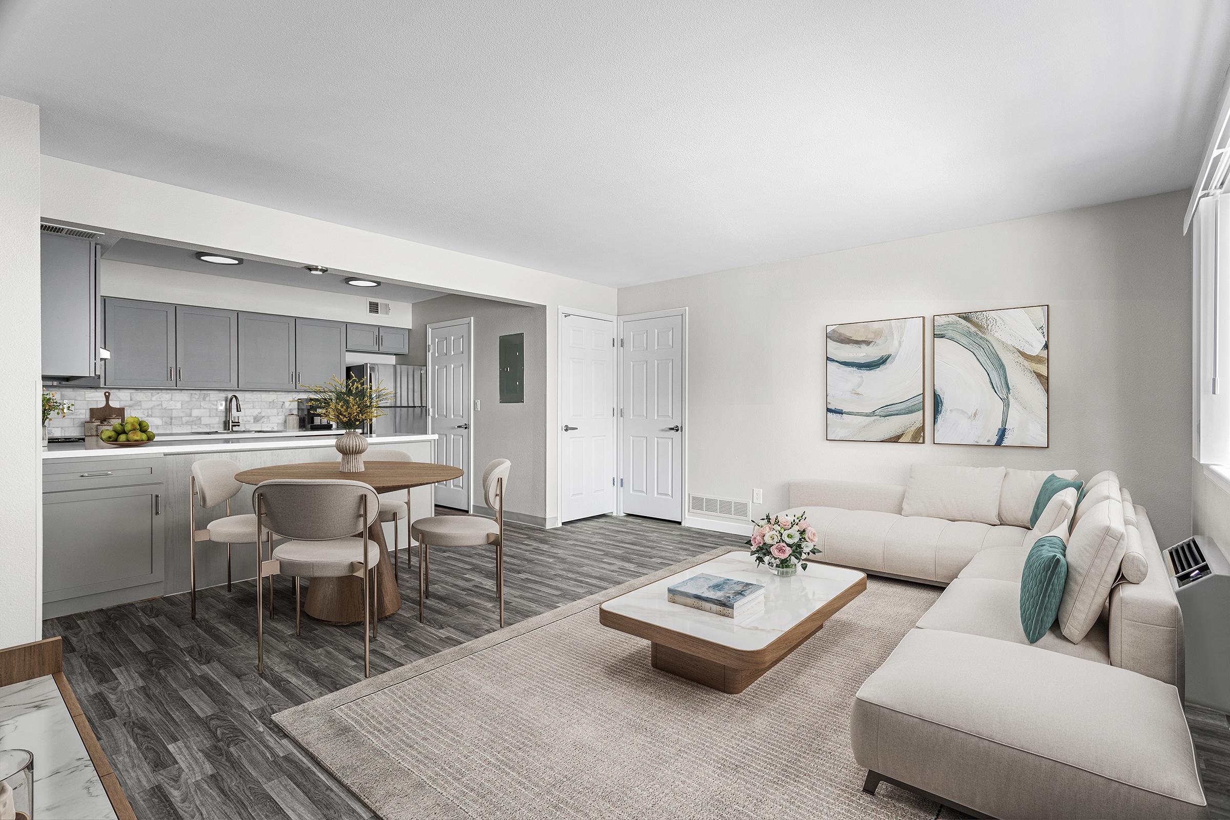 A modern and stylish living room with a light color palette. It features a comfortable beige sectional sofa, a wooden coffee table, and a round dining table with chairs. The kitchen is partially visible with gray cabinets and a marble backsplash. Two abstract artworks are mounted on the wall, adding a touch of decor.