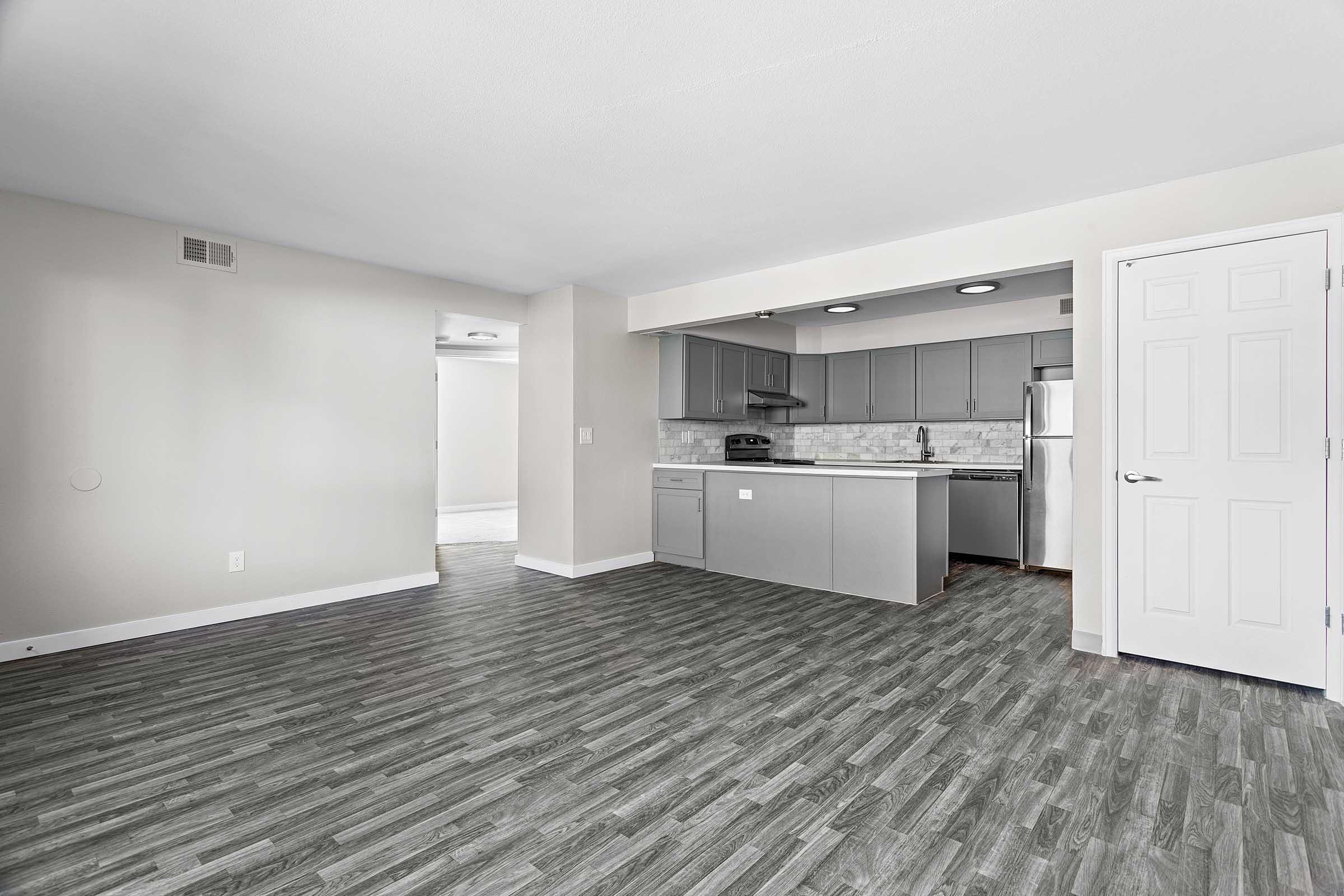 A spacious, modern kitchen and living area featuring gray cabinets, stainless steel appliances, and a light-colored tile backsplash. The flooring is a wood-like vinyl with gray tones, and the walls are painted in a soft beige. Bright light enters through the open space, enhancing the inviting atmosphere.