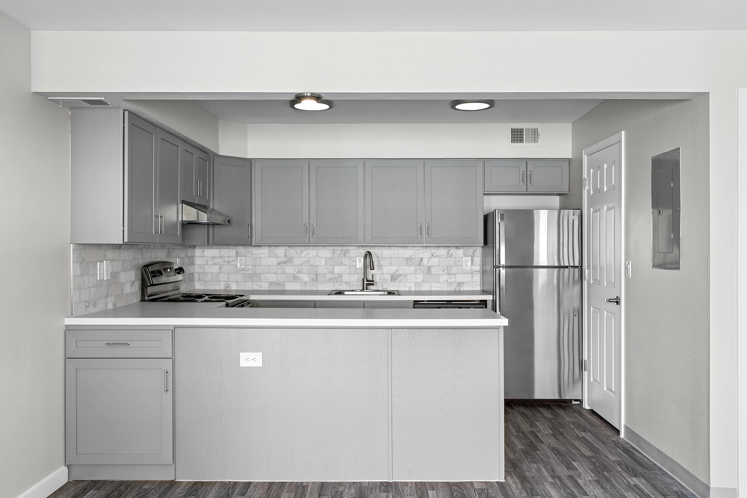Modern kitchen featuring gray cabinets, a marble backsplash, stainless steel appliances, and a spacious countertop. The area is well-lit with recessed lighting and has an open design that connects to an adjacent room. The floor is dark wood-like laminate, enhancing the contemporary aesthetic.