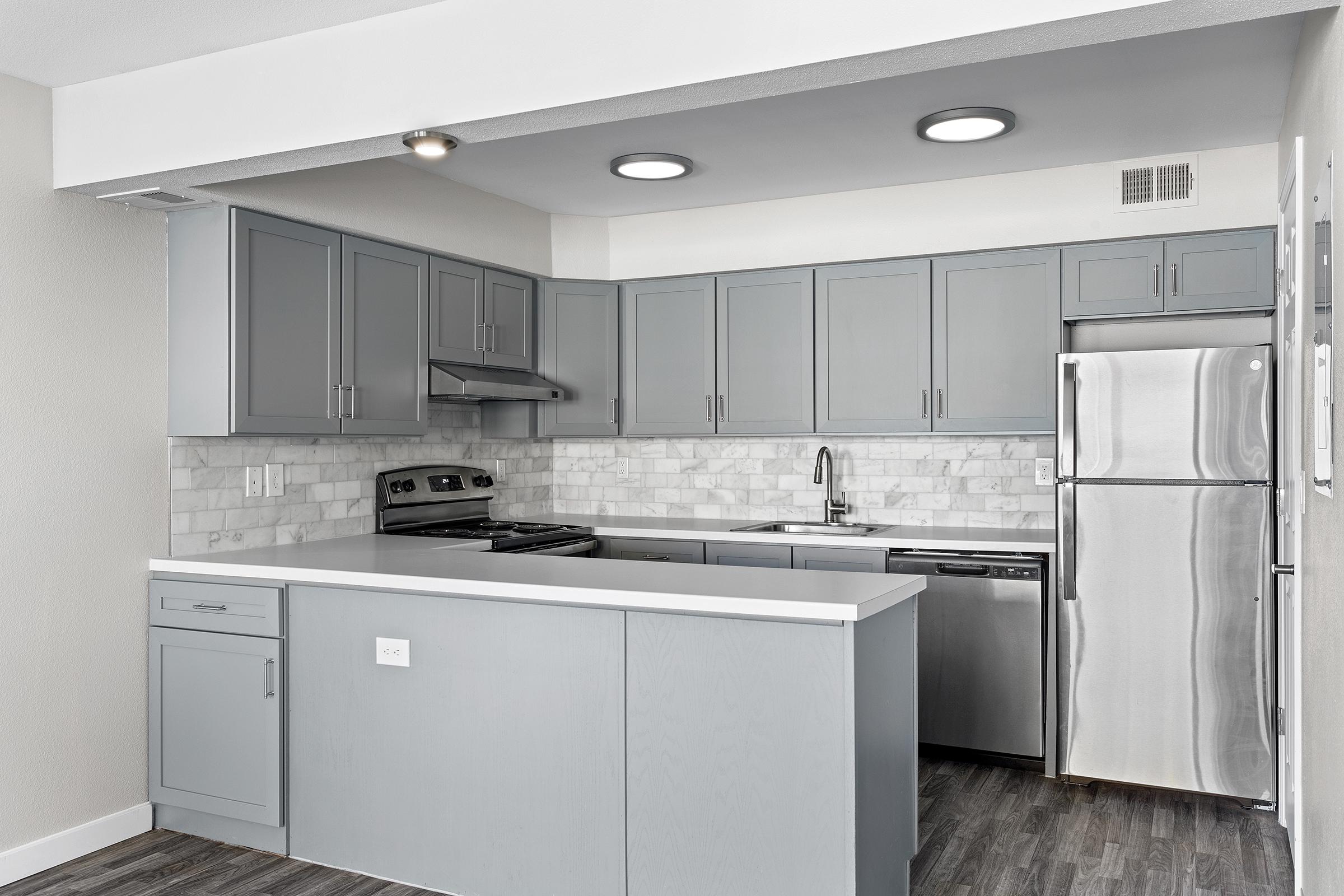 Modern kitchen featuring light gray cabinets, a stainless steel refrigerator, and an electric stove. The countertop is white, complemented by a marble backsplash. The space is well-lit with recessed ceiling lights and has a sleek, contemporary design.