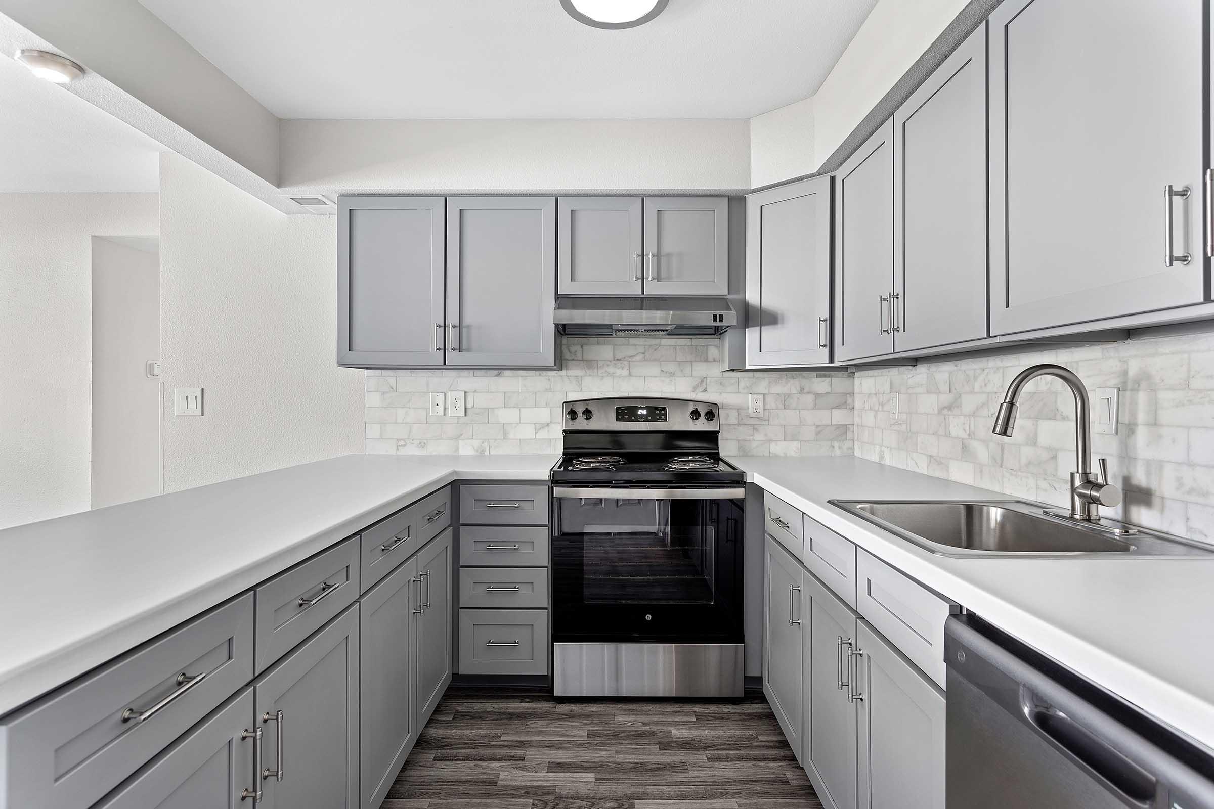 Modern kitchen with gray cabinets, a stainless steel oven, and a sleek countertop. The backsplash features light-colored tiles, and a stainless steel sink is installed. The flooring has a wood-like appearance, creating a contemporary and inviting atmosphere. Bright lighting enhances the overall aesthetic.