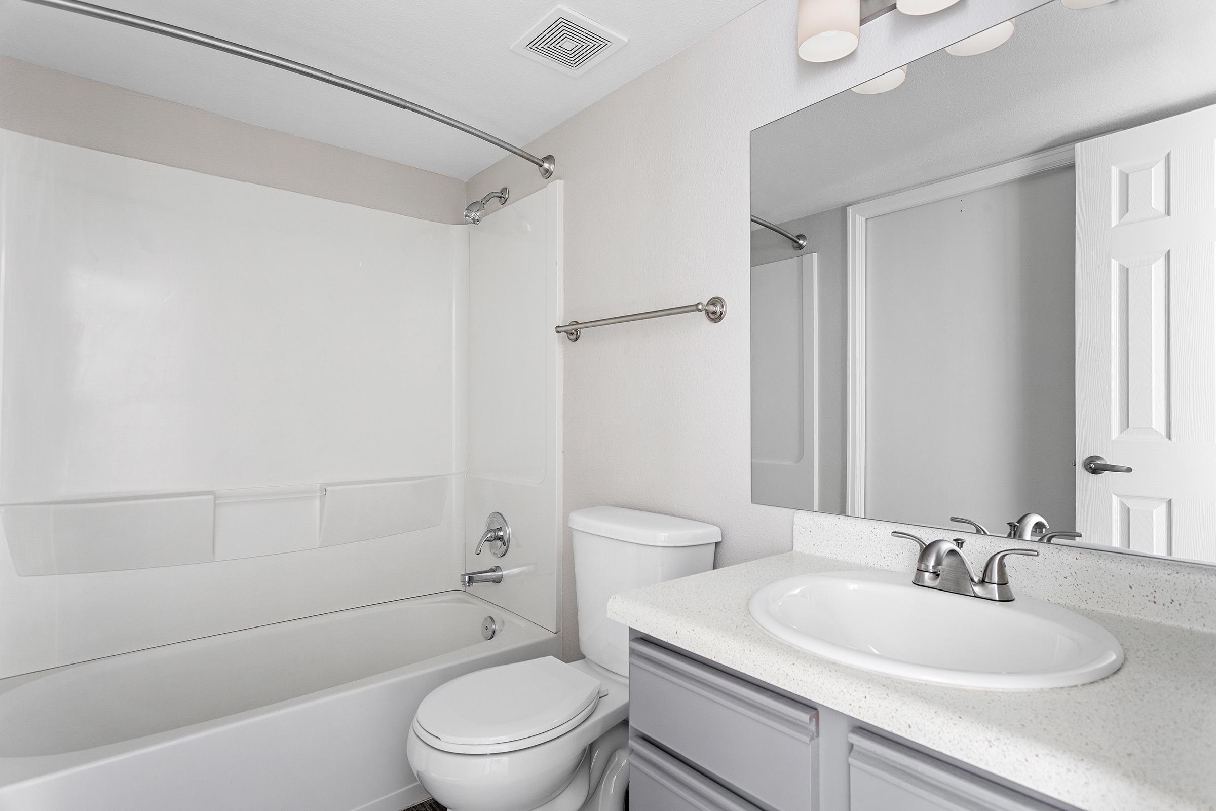 A modern bathroom featuring a white tub with a shower curtain, a toilet, and a vanity with a sink and chrome faucet. The walls are a light color, and there is a large mirror above the sink. A towel bar is mounted on the wall, and there is a door leading to another room. Natural light is present.