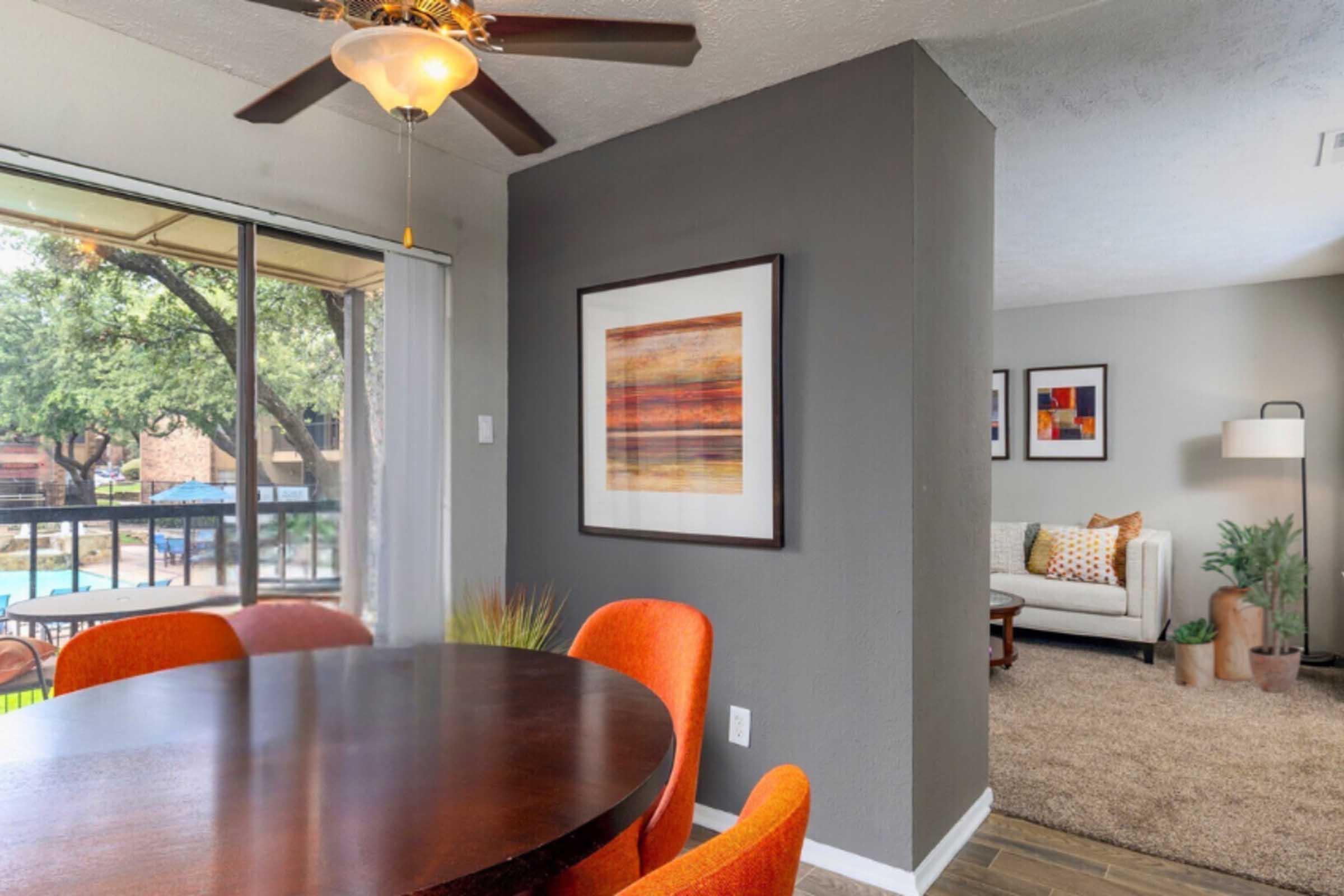 A modern dining area featuring a round wooden table surrounded by orange chairs, a ceiling fan, and large windows with natural light. A gray accent wall with framed artwork complements the space, while a cozy living area with a sofa and decorative plants is visible in the background.