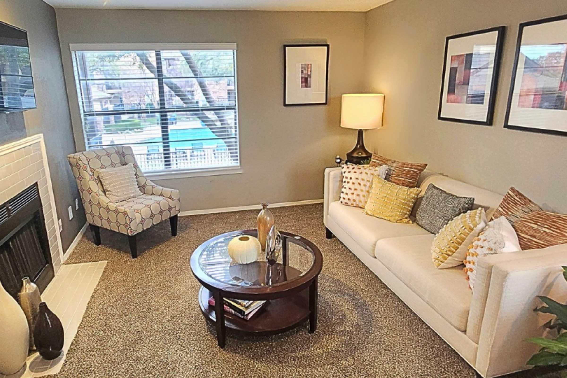 A cozy living room featuring a white sofa adorned with colorful pillows, a round coffee table with decorative items, and a patterned armchair. The walls are painted in a soft color, with framed artwork, and a large window provides a view of an outdoor pool area.