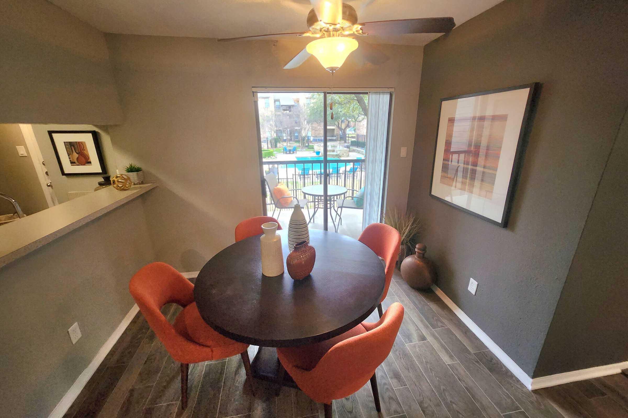 A modern dining area featuring a round table with black surface and four orange chairs. A decorative vase sits on the table. In the background, there is a sliding glass door leading to a pool area, with a view of trees and outdoor seating. The walls are painted in dark tones, creating a cozy ambiance.
