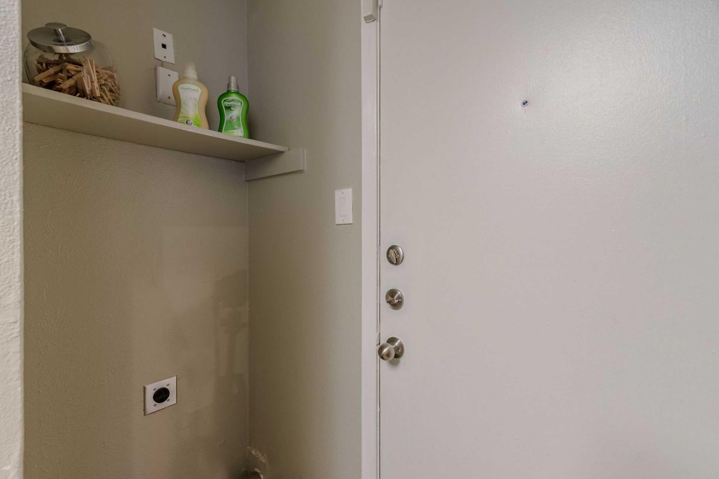 A view of a simple entryway with a closed white door, a shelf on the left holding bottles of cleaning products, and a small container of wooden sticks. There is a wall near the door with an electrical outlet. The overall color scheme is neutral and minimalistic.