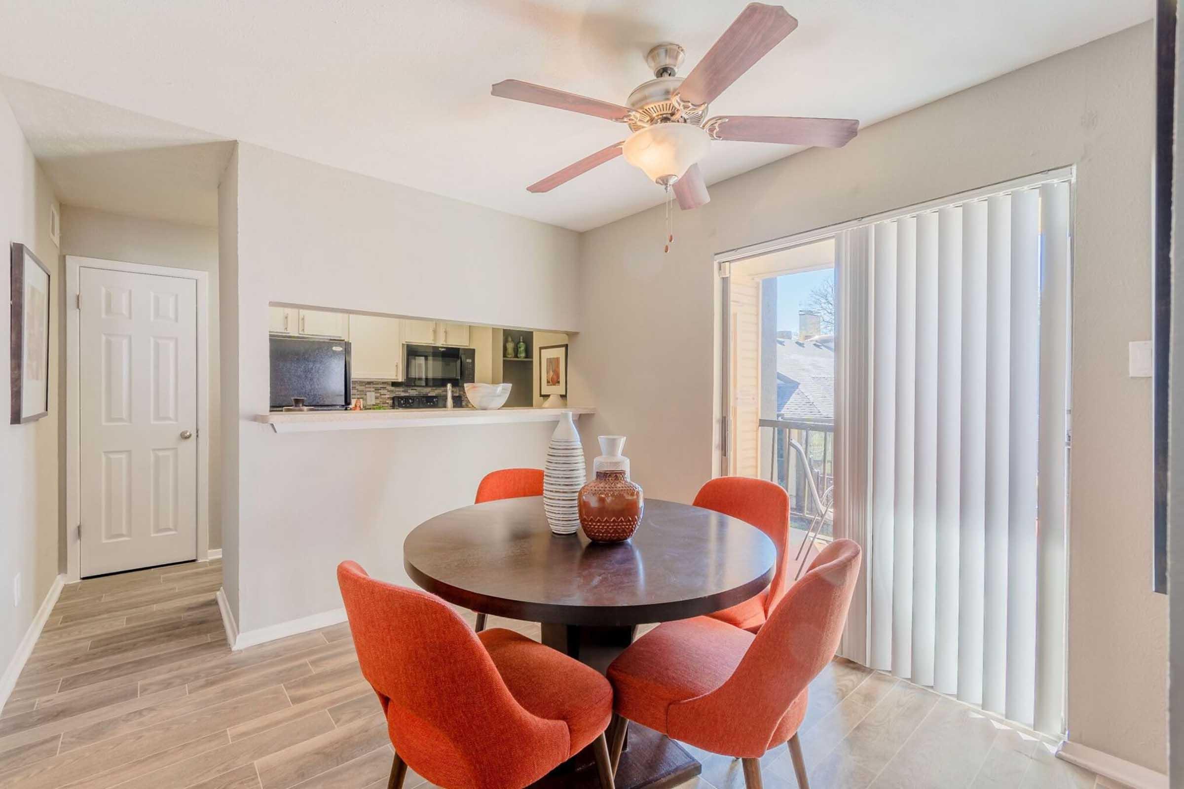 A cozy dining area featuring a round wooden table with four orange chairs, a decorative vase in the center, and a ceiling fan overhead. Large sliding glass doors lead to a balcony, while a kitchen area is visible in the background, complete with a microwave and small TV. Bright, neutral walls enhance the inviting atmosphere.