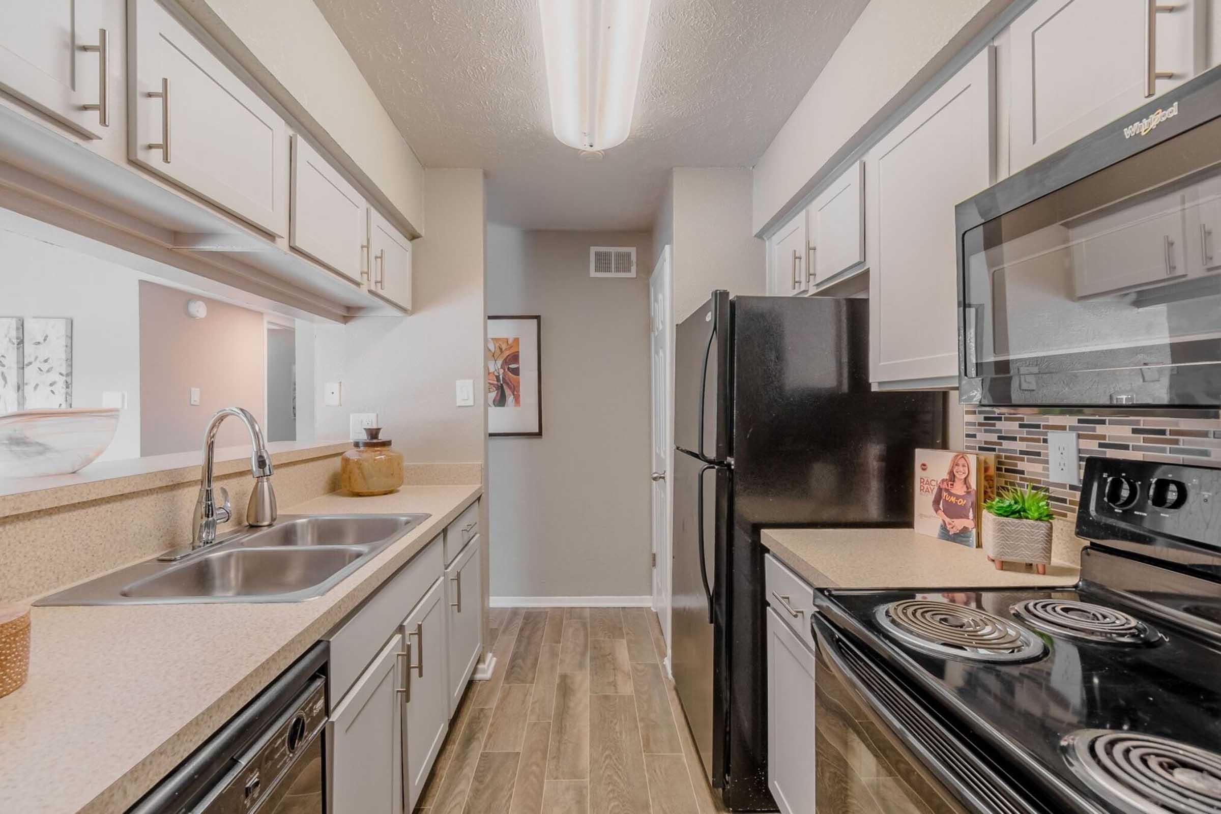 Modern kitchen featuring a long countertop with a double sink, stainless steel appliances, including a stove and refrigerator. Light-colored cabinets line the walls, and there's a decorative backsplash. The space is well-lit with overhead lighting and has a neutral color palette with wooden flooring.