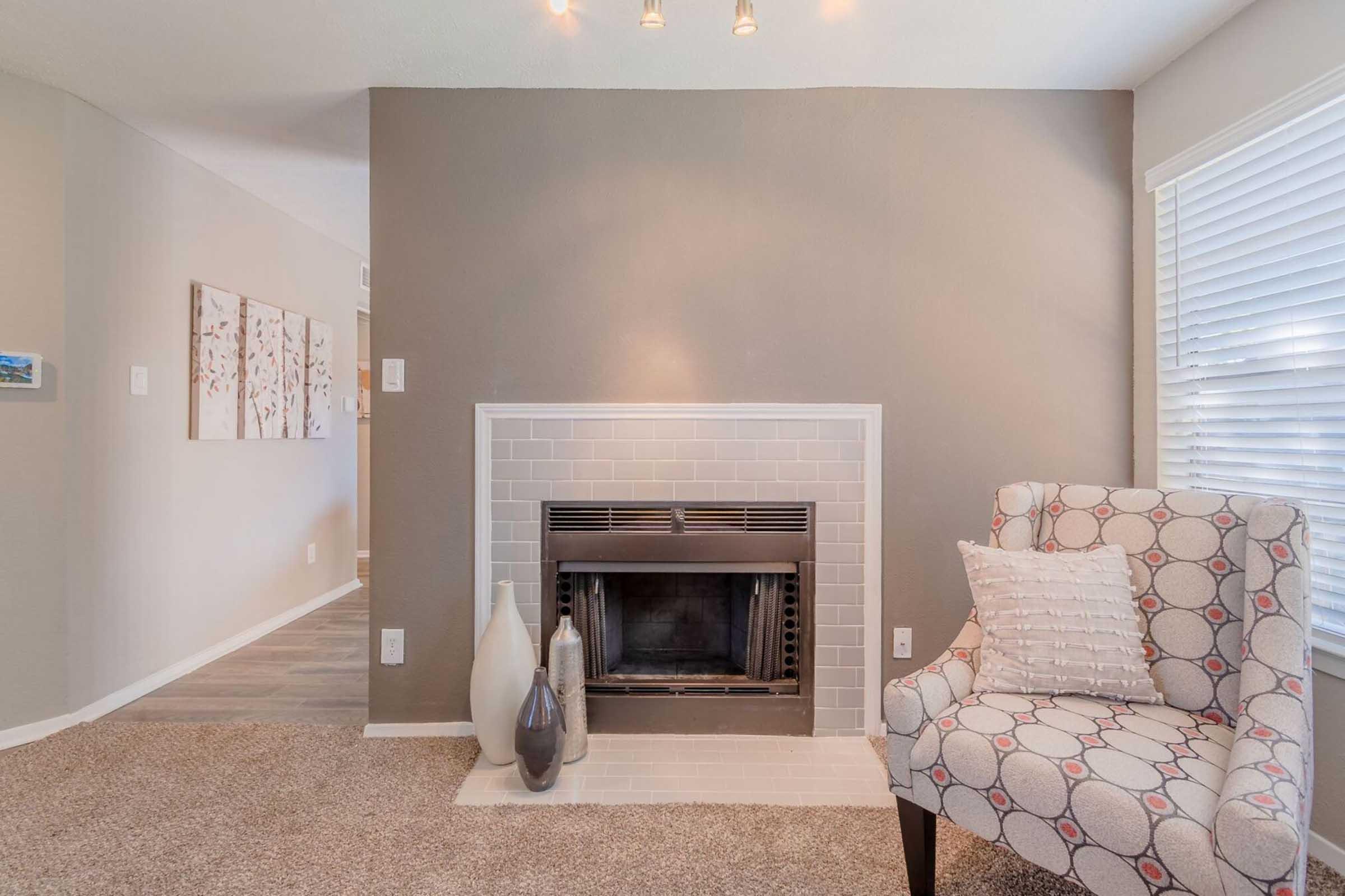 A cozy interior featuring a decorative chair next to a modern fireplace with a grey tile surround. The wall is painted in neutral tones, complemented by a light-colored carpet. A small vase sits on the floor beside the fireplace. Natural light streams in through the window with blinds.