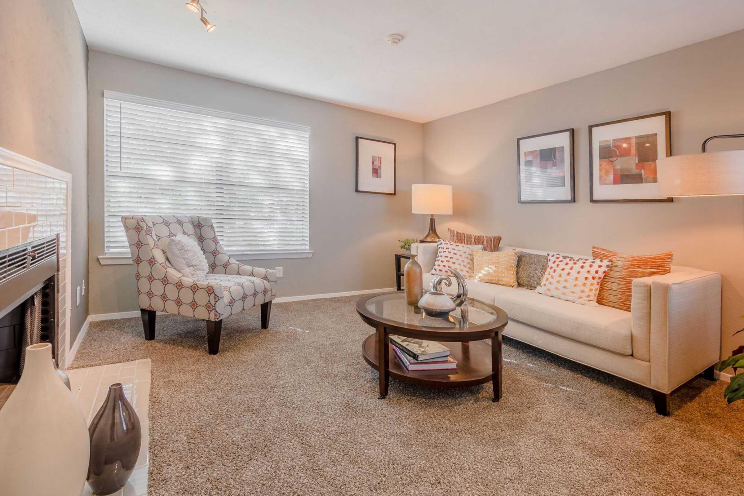 A cozy living room featuring a light-colored couch with decorative pillows, a round coffee table with magazines, and a stylish chair. The walls are painted in neutral tones, and there are framed artworks. Natural light filters through a window with blinds, creating a warm atmosphere. A soft carpet completes the space.