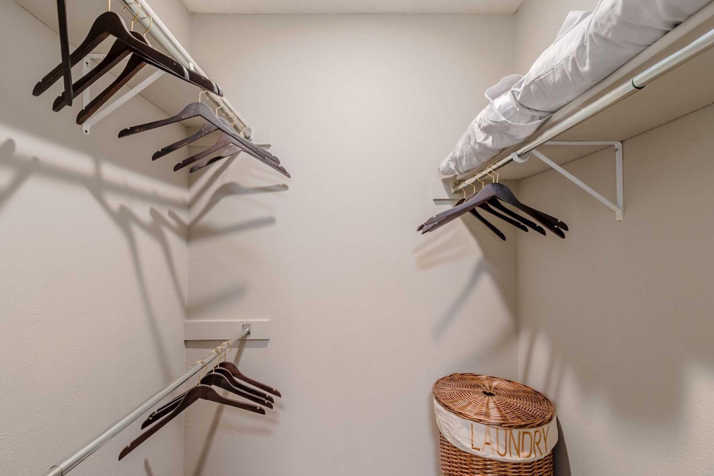 A neatly organized closet featuring multiple wooden hangers on white metal rods, a neatly stacked blanket on a shelf above, and a wicker laundry basket labeled "LAUNDRY" placed on the floor. The walls are light-colored, contributing to a clean and spacious appearance.