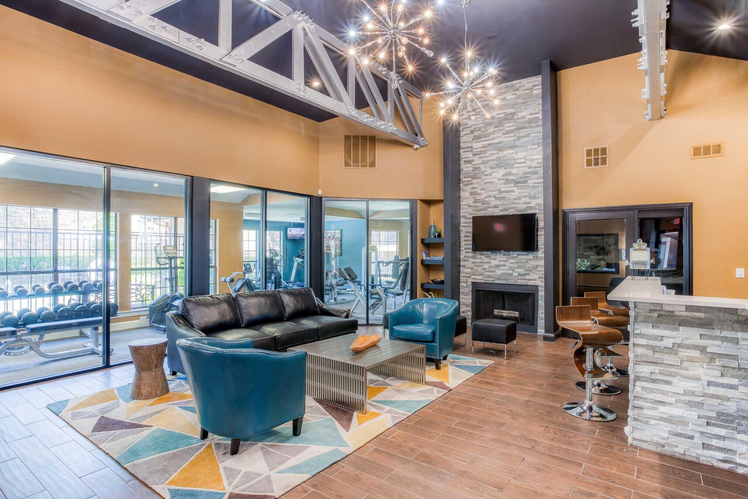 Modern lounge area featuring a black leather sofa, turquoise accent chairs, and a stone fireplace. Large windows provide a view of fitness equipment in a gym area. The space is decorated with a geometric area rug and stylish light fixtures, creating a welcoming atmosphere.