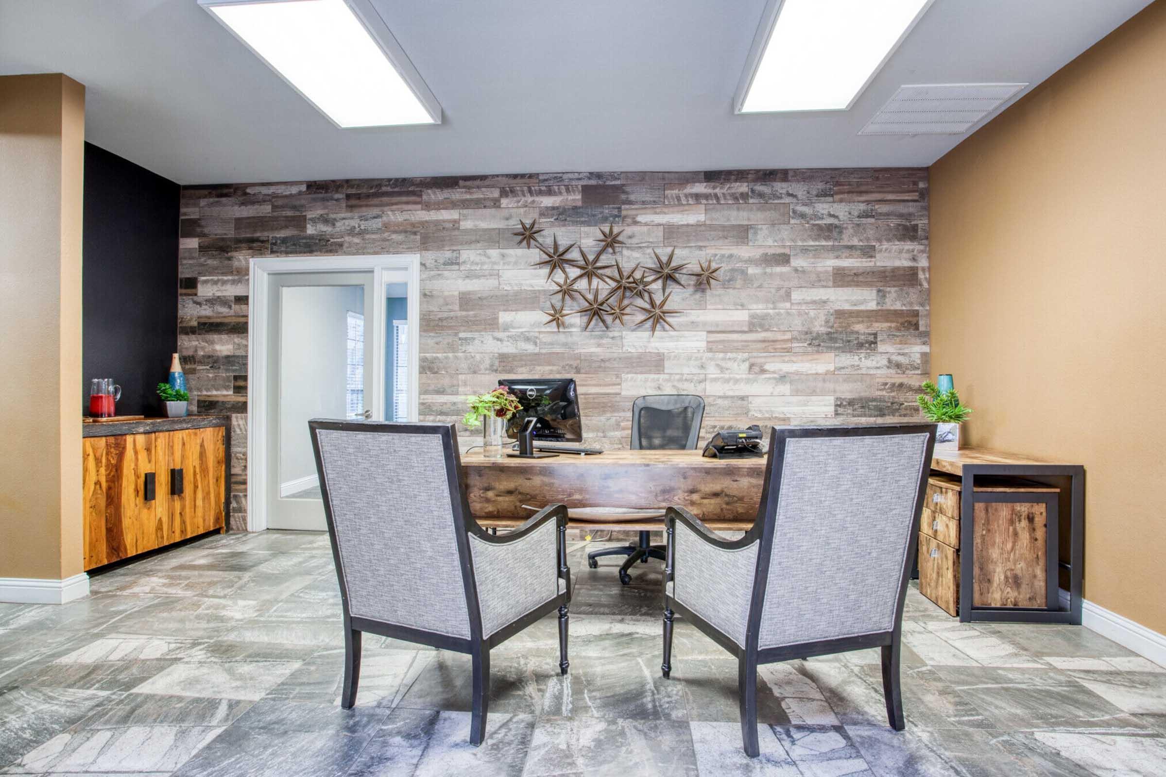 A modern office space featuring a wooden desk with a black ergonomic chair, two grey upholstered chairs, and a decorative wall with star-shaped accents. The walls are a warm beige and a dark finish, with a stone-like patterned floor and some greenery for a fresh touch. Bright overhead lights illuminate the room.