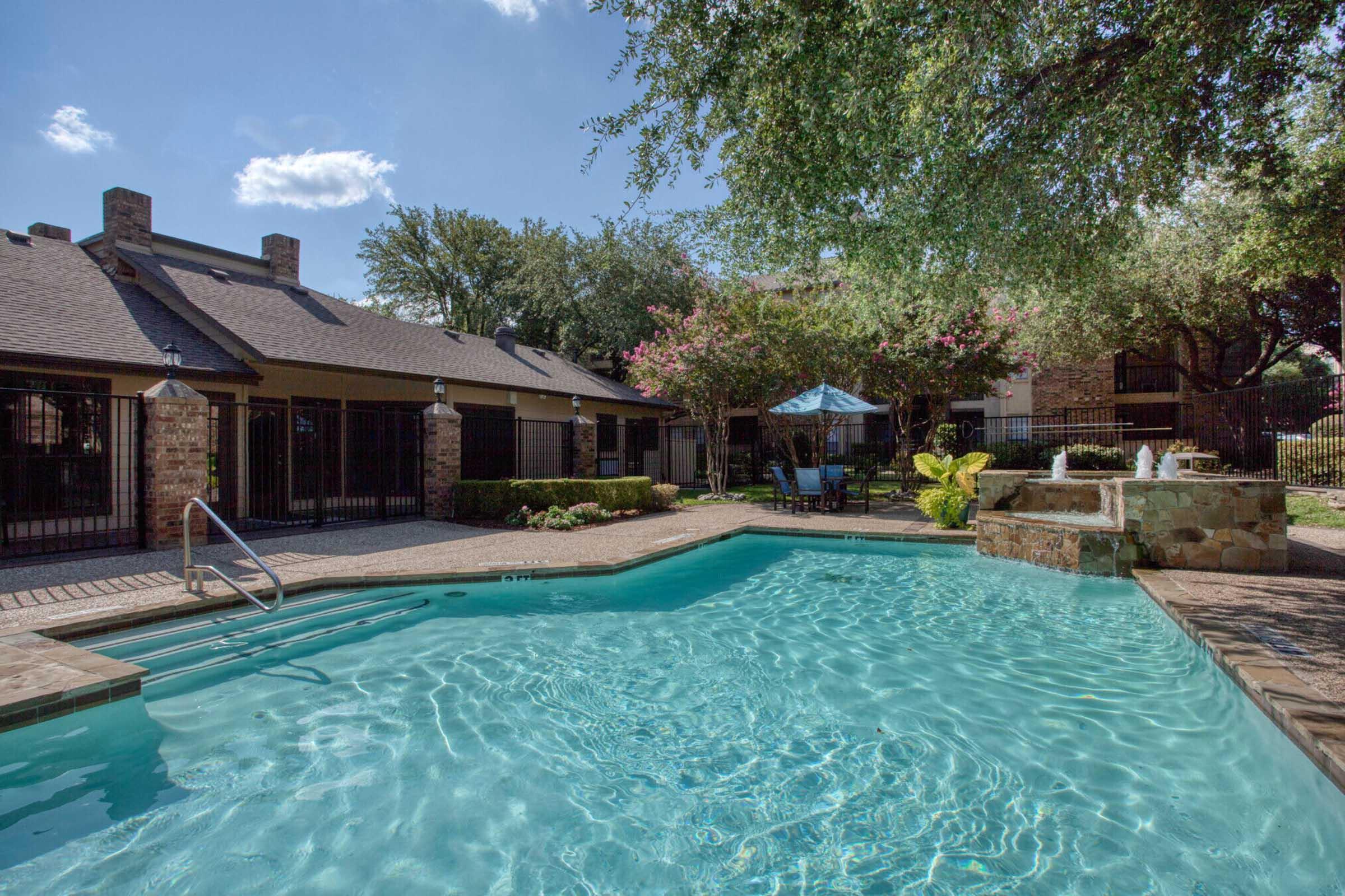 A serene swimming pool surrounded by landscaped greenery and stone pathways. There are lounge chairs nearby, a shaded seating area with a blue umbrella, and decorative plants. The background features a brick building and blue sky with a few fluffy clouds, creating a relaxing outdoor atmosphere.