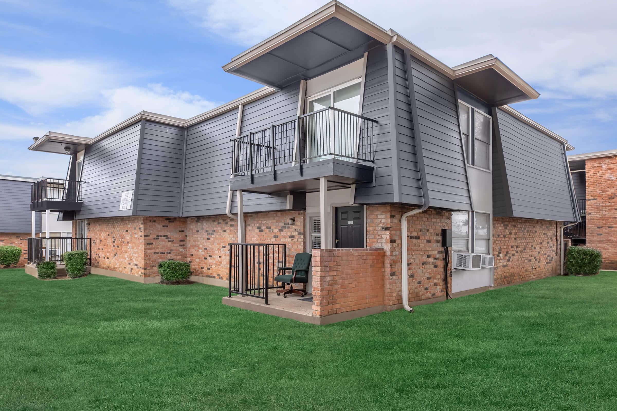 A two-story apartment building featuring modern gray siding and brick accents. The structure has several balconies with metal railings, and air conditioning units are visible. Well-maintained green grass surrounds the building, creating a tidy outdoor space. The sky is partly cloudy.