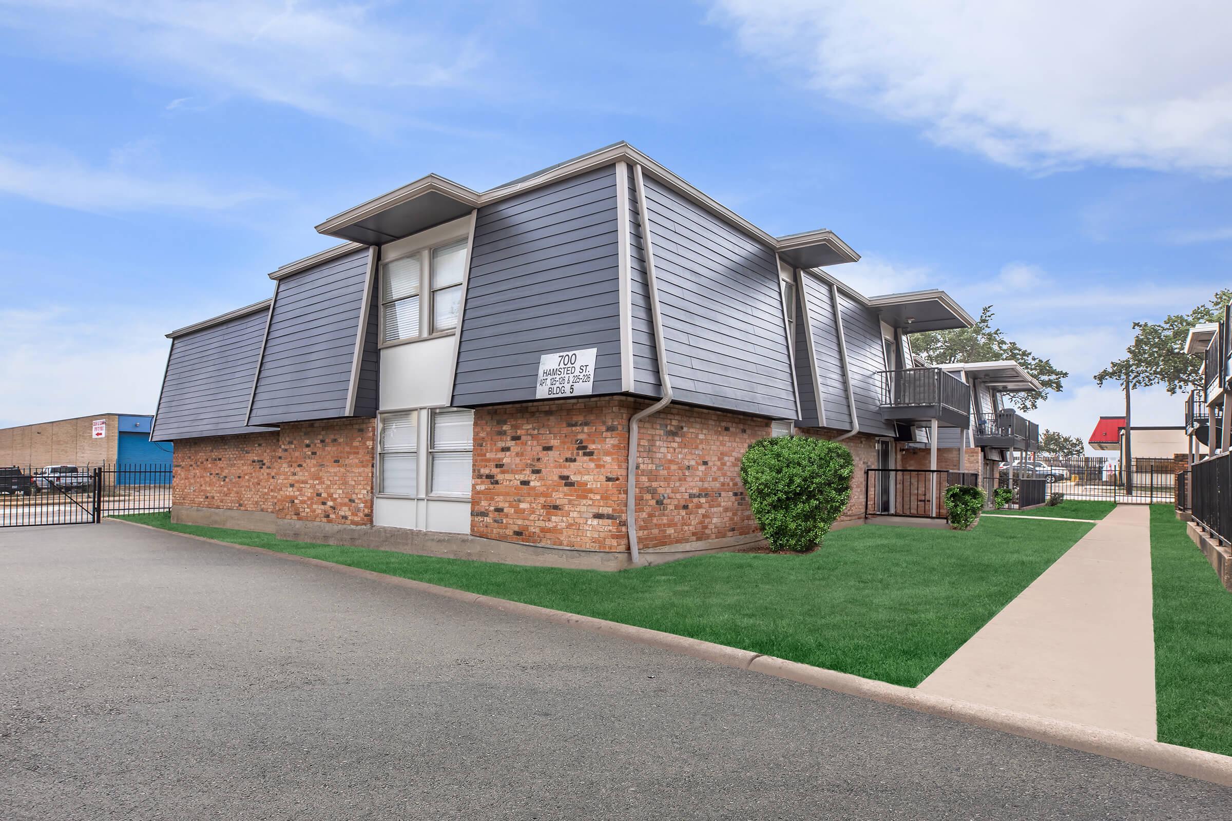 Exterior view of a two-story apartment building featuring a combination of gray siding and brick. The building has balconies on the second floor and is situated on a landscaped area with grass. A paved walkway runs alongside the structure, with a parking area visible in the background.