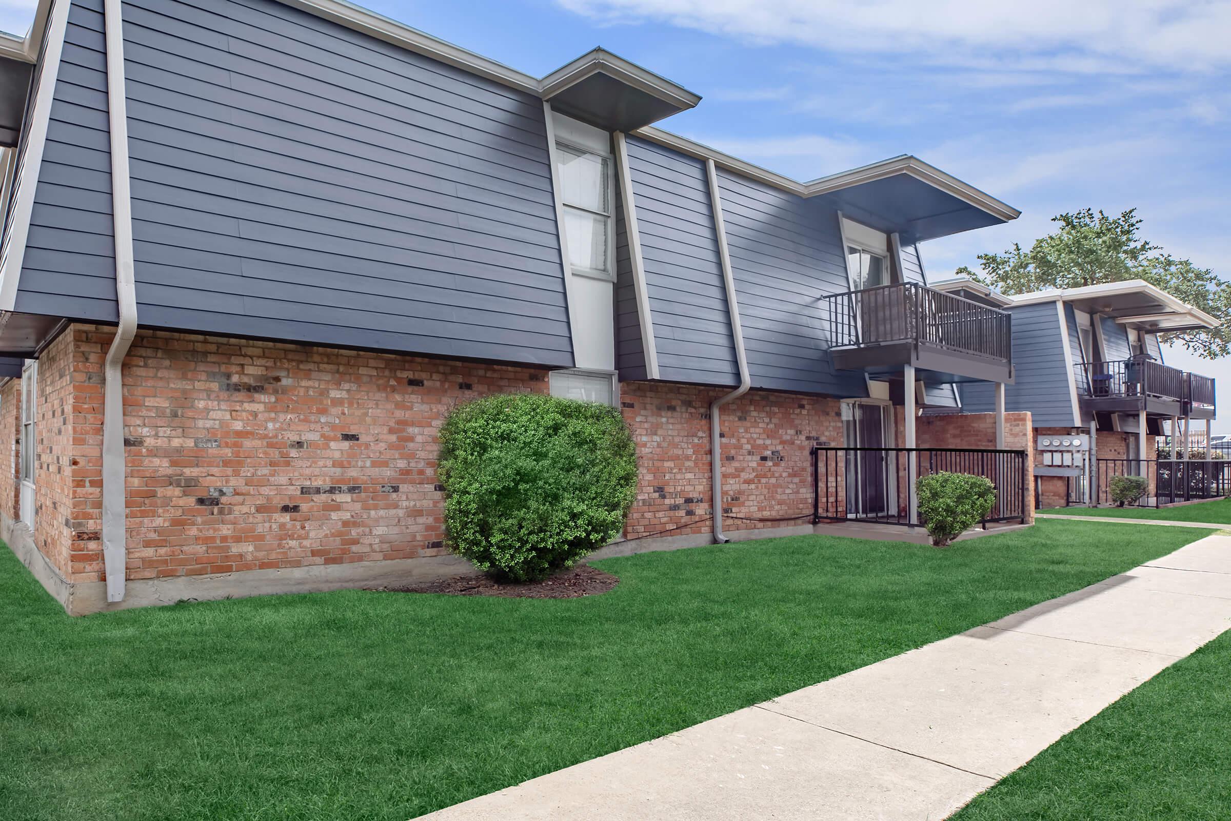 A view of a modern two-story apartment building featuring a combination of gray siding and brick. The structure includes balconies on the upper level, surrounded by well-maintained green lawn and a concrete pathway leading to the entrance. Clear blue sky in the background.