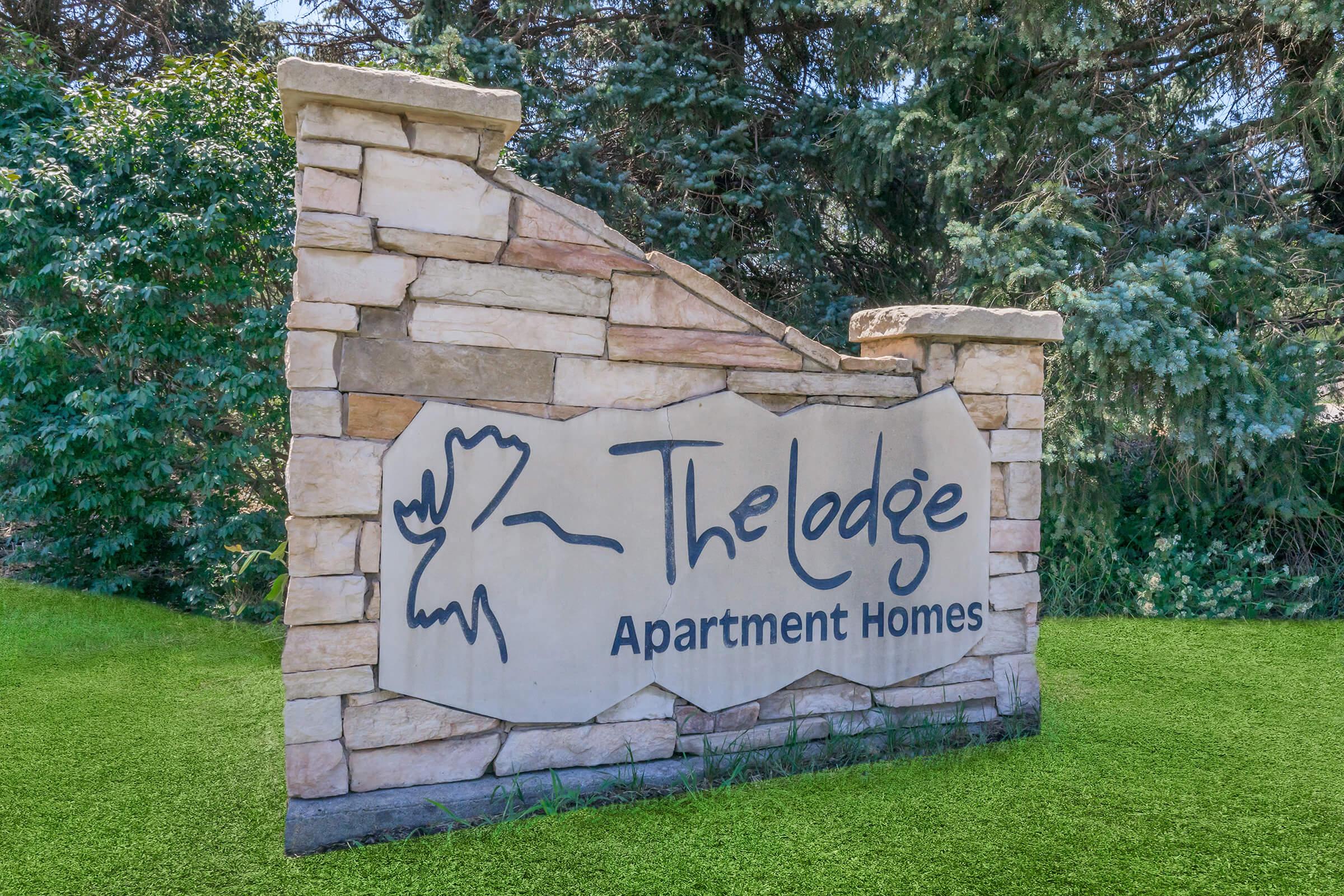 Sign at The Lodge Apartment Homes featuring a stone structure with a stylized logo and text. Surrounded by greenery, the sign presents a welcoming entrance to the apartment complex.
