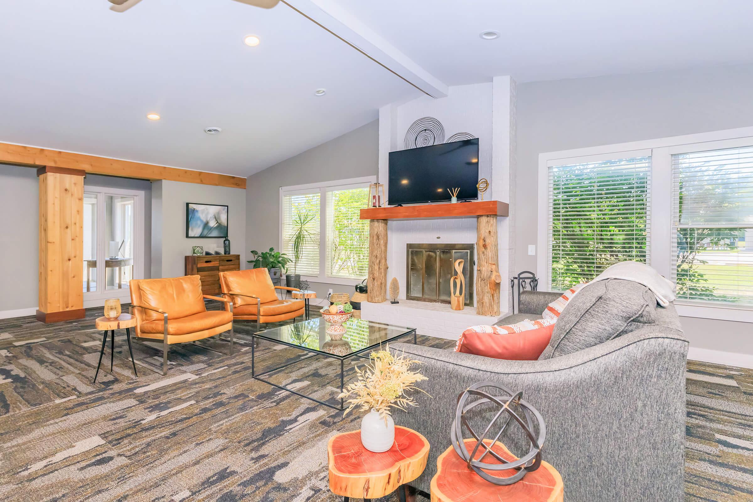 A modern living room with large windows letting in natural light. It features a gray sofa, two orange armchairs, a glass coffee table, and a stylish fireplace with a mounted TV above it. Decorative elements include a wooden beam and small plants, creating a cozy and inviting atmosphere.