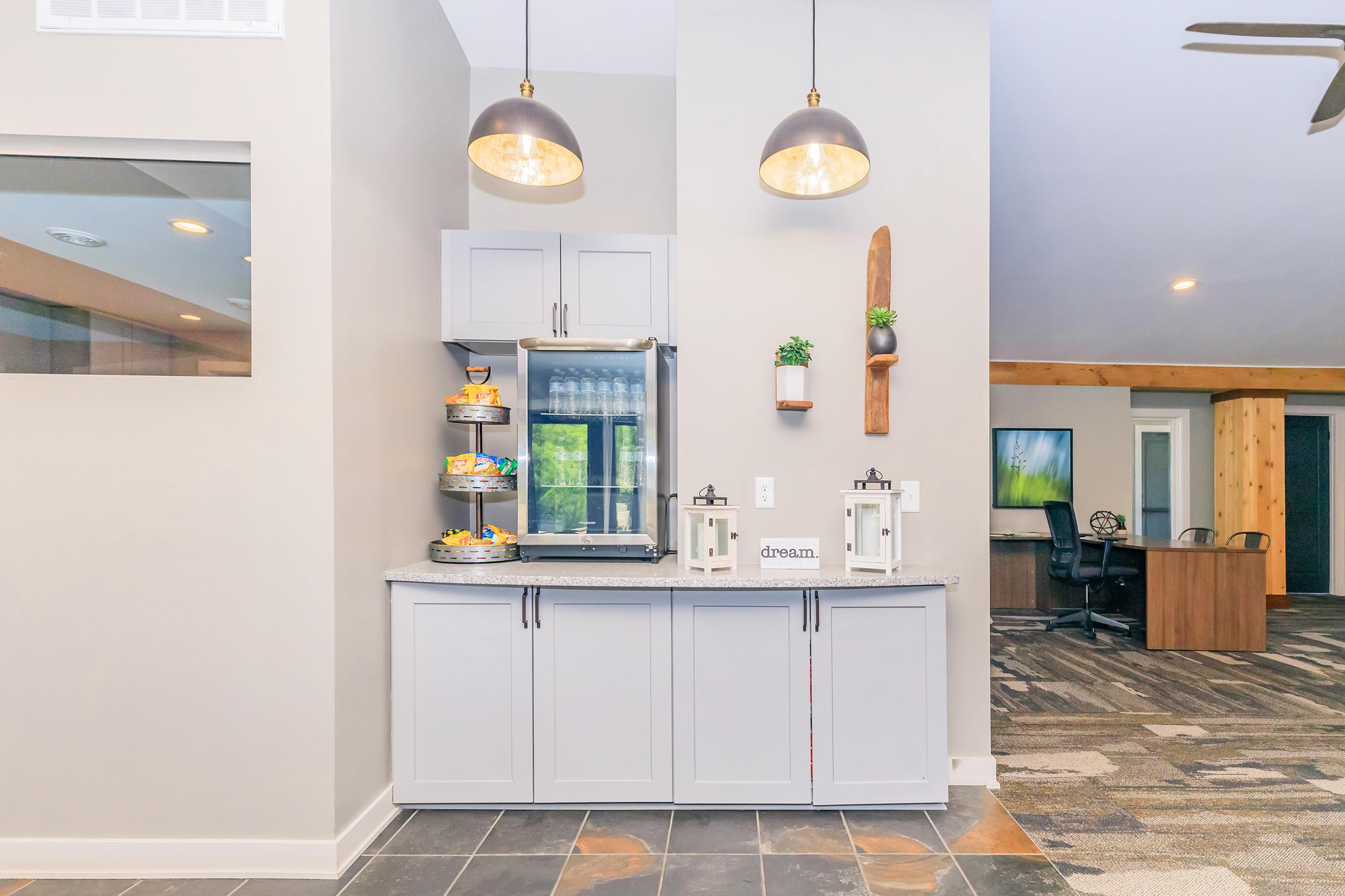 A modern interior space featuring a light gray cabinet with a countertop, a glass display stand filled with snacks, and decorative items like potted plants and lanterns. Pendant lights hang above, and soft flooring leads to a connected workspace with wooden accents.