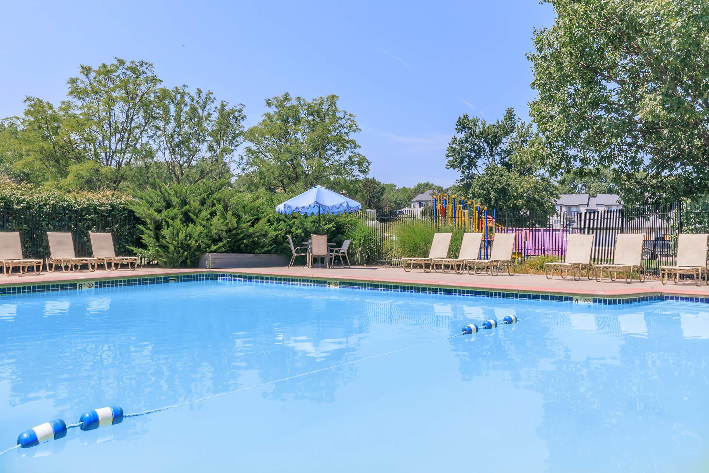 A clear blue swimming pool surrounded by lounge chairs and greenery. In the background, there is a colorful play structure and a shaded seating area with an umbrella. The sky is bright and sunny, indicating a pleasant outdoor atmosphere.