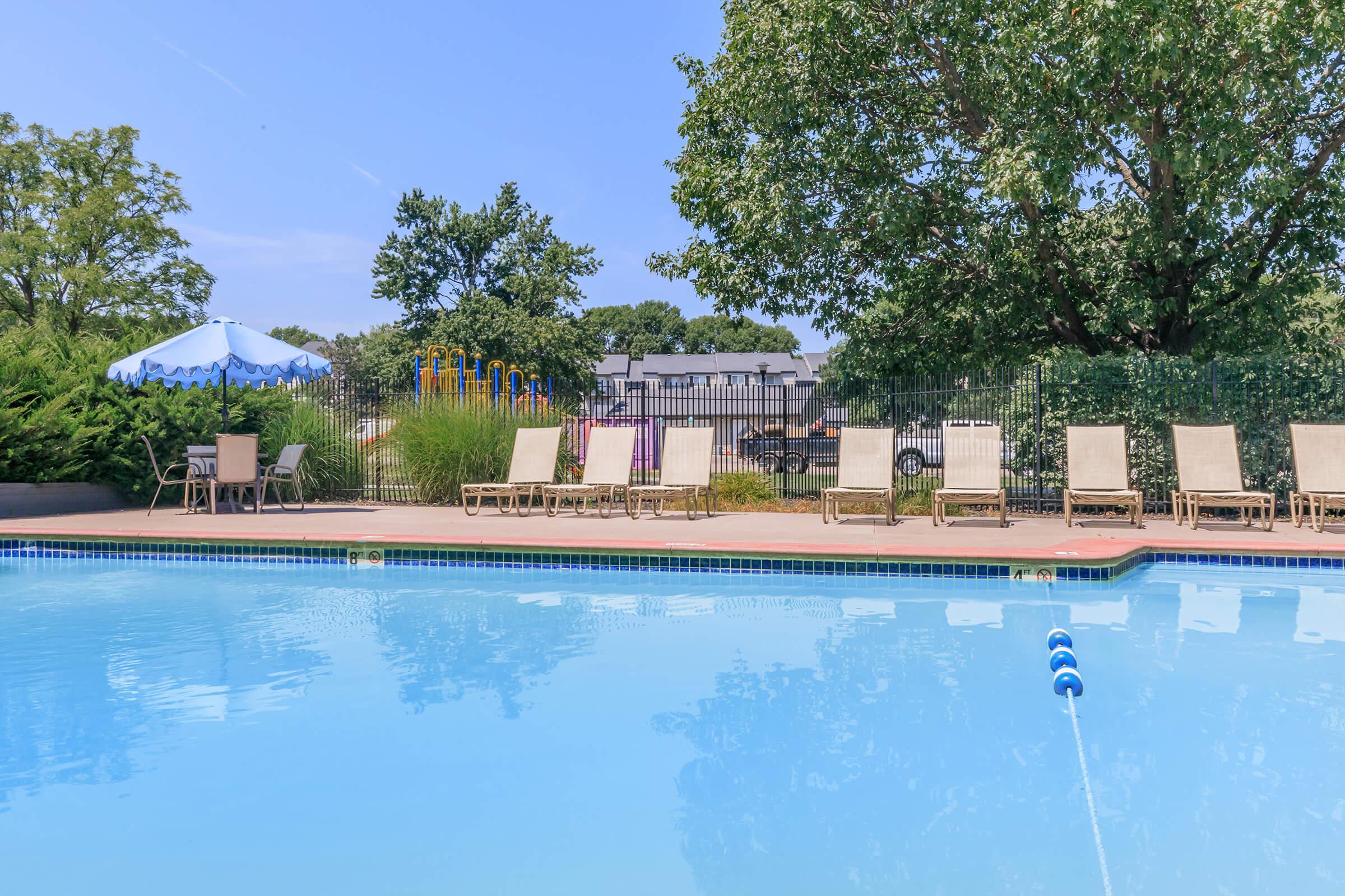 A clear swimming pool surrounded by lounge chairs, with a blue and white umbrella in the background. The pool's surface reflects the sky and trees, while a fenced area with playground equipment is visible in the distance.
