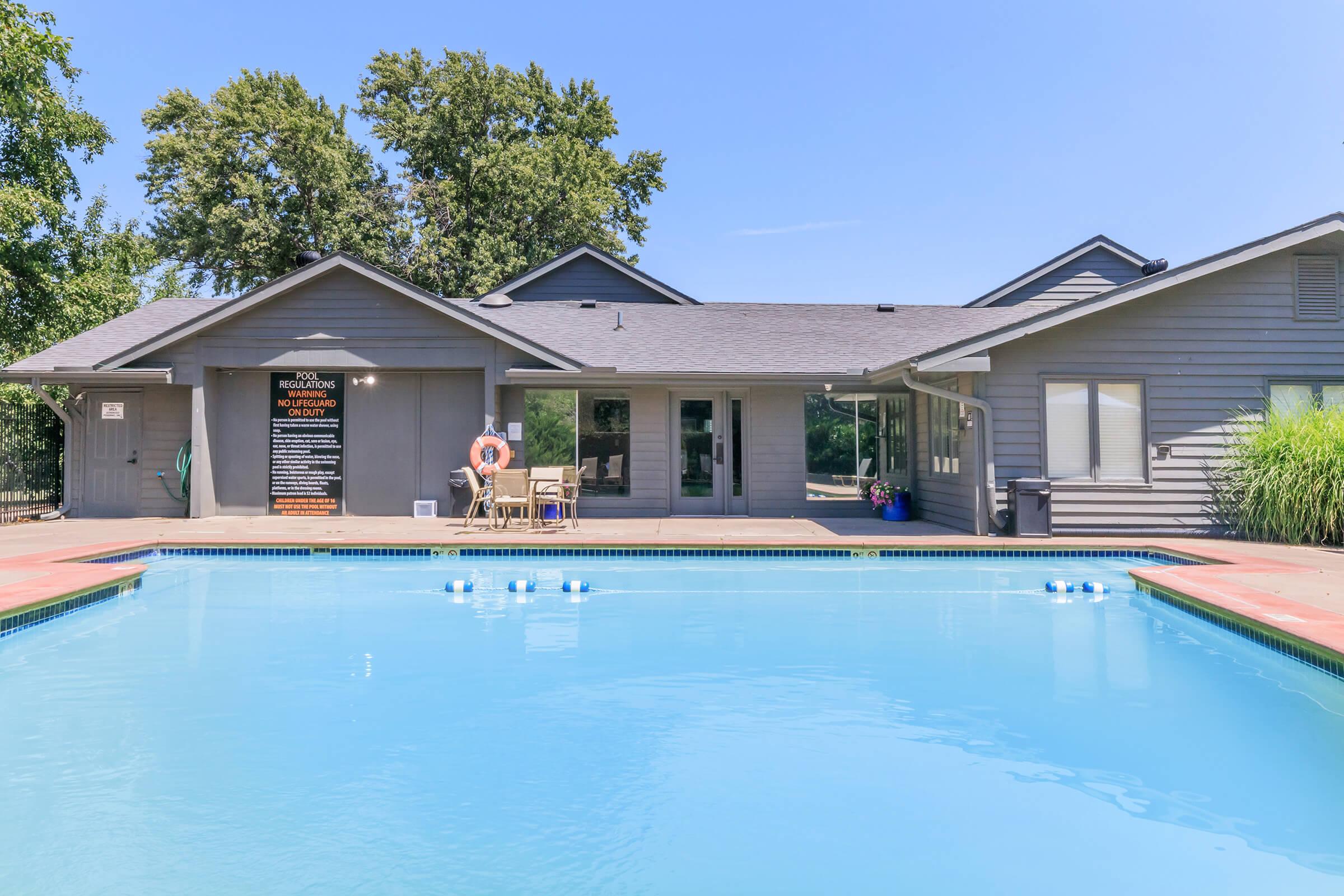 A bright and inviting outdoor swimming pool area surrounded by a wooden deck. In the background, there is a modern building with large windows and a sign. Lounge chairs are placed near the pool, and there are trees and greenery in the surrounding landscape. Clear blue skies overhead complete the scene.