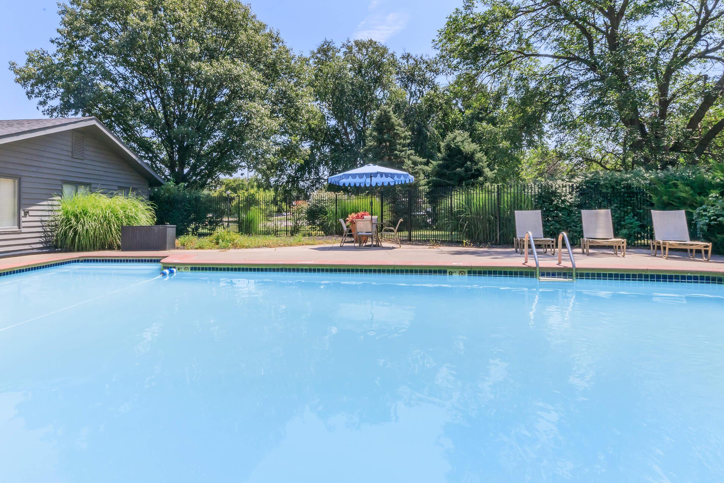 A clear blue swimming pool surrounded by lush greenery. In the background, there's a patio with a striped umbrella and outdoor chairs. The scene is bright and sunny, showcasing a relaxed outdoor space ideal for leisure and enjoyment.