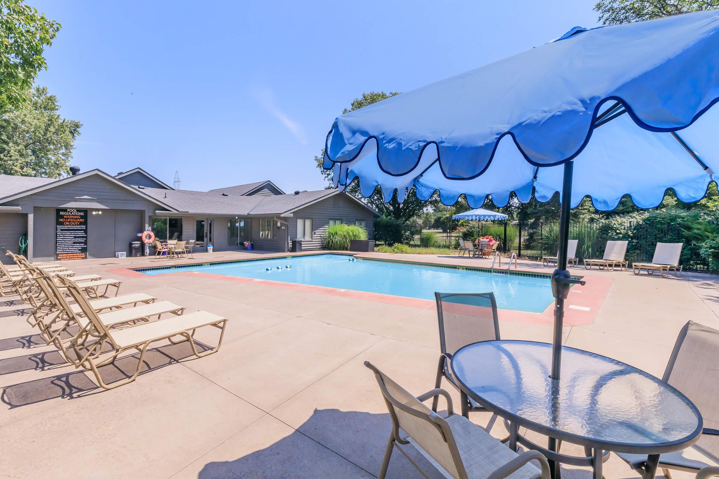 A swimming pool surrounded by lounge chairs and tables. A blue umbrella provides shade over a table. In the background, there are several houses and lush greenery. The sky is clear and blue, indicating a sunny day. The pool area offers a relaxing outdoor space for leisure.