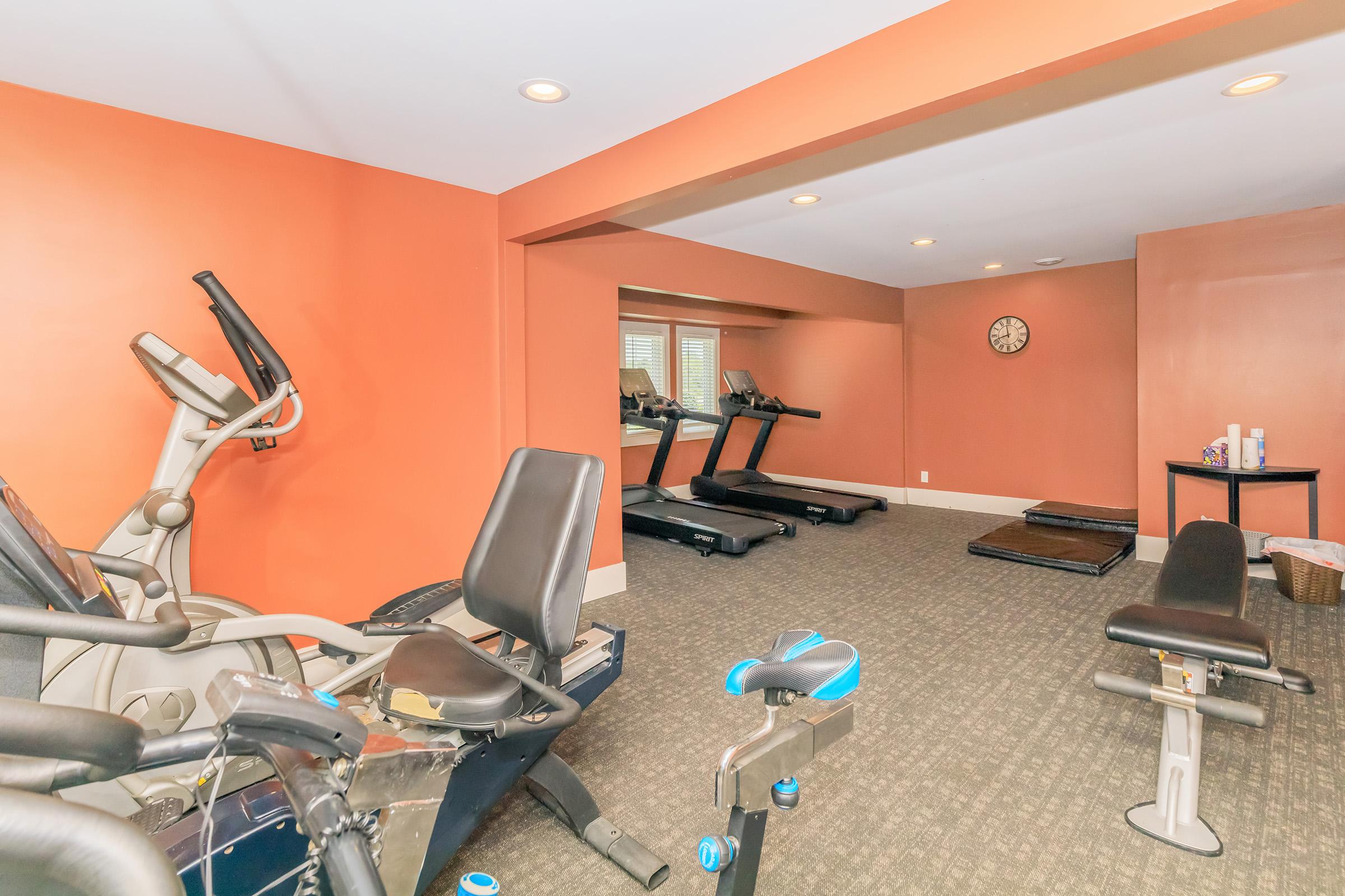 A well-lit home gym featuring exercise equipment including treadmills, an elliptical machine, and a weight bench. The walls are painted orange, and there is a mirror on one side reflecting the workout area. A clock is mounted on the wall, and a small table holds fitness supplies.