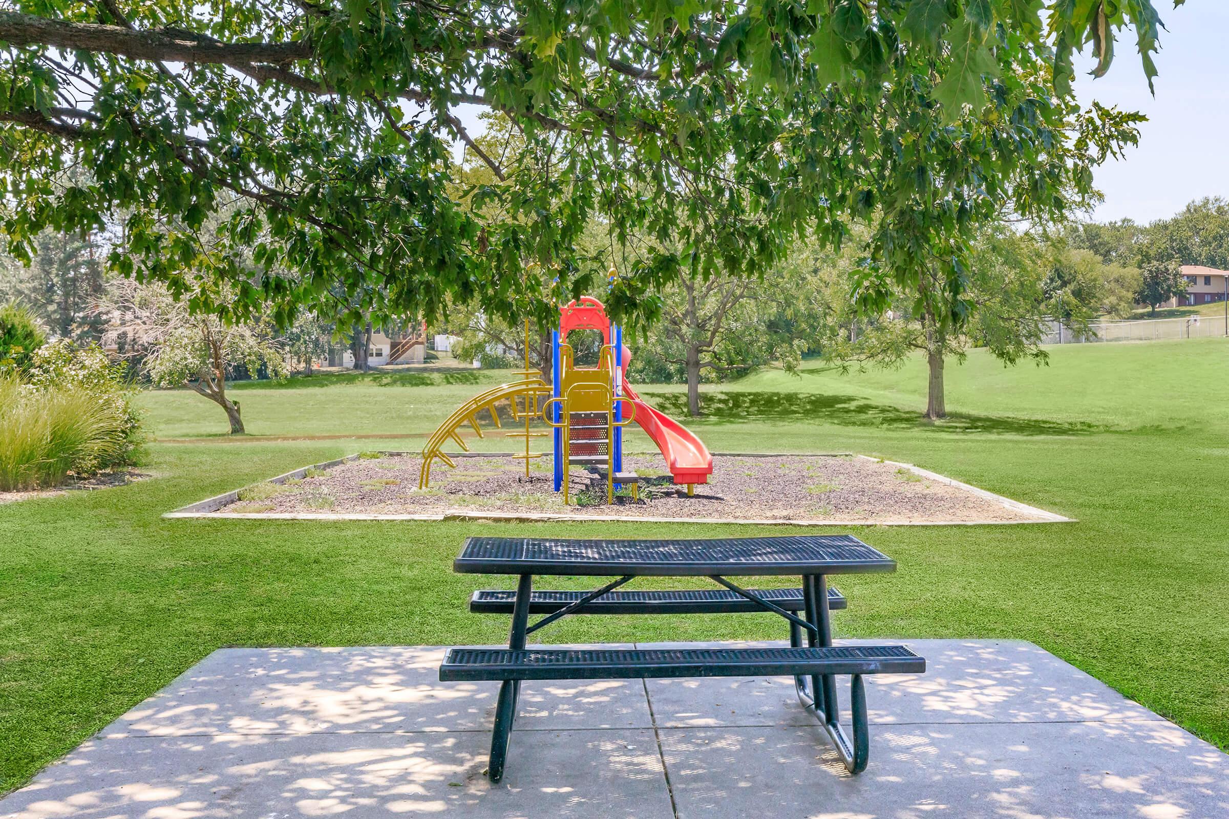 A playground featuring a colorful slide and climbing structure is set within a grassy park. In the foreground, there's a black picnic table on a concrete slab, surrounded by trees and an open green space. The scene is bright and inviting, perfect for family outings or children's playtime.