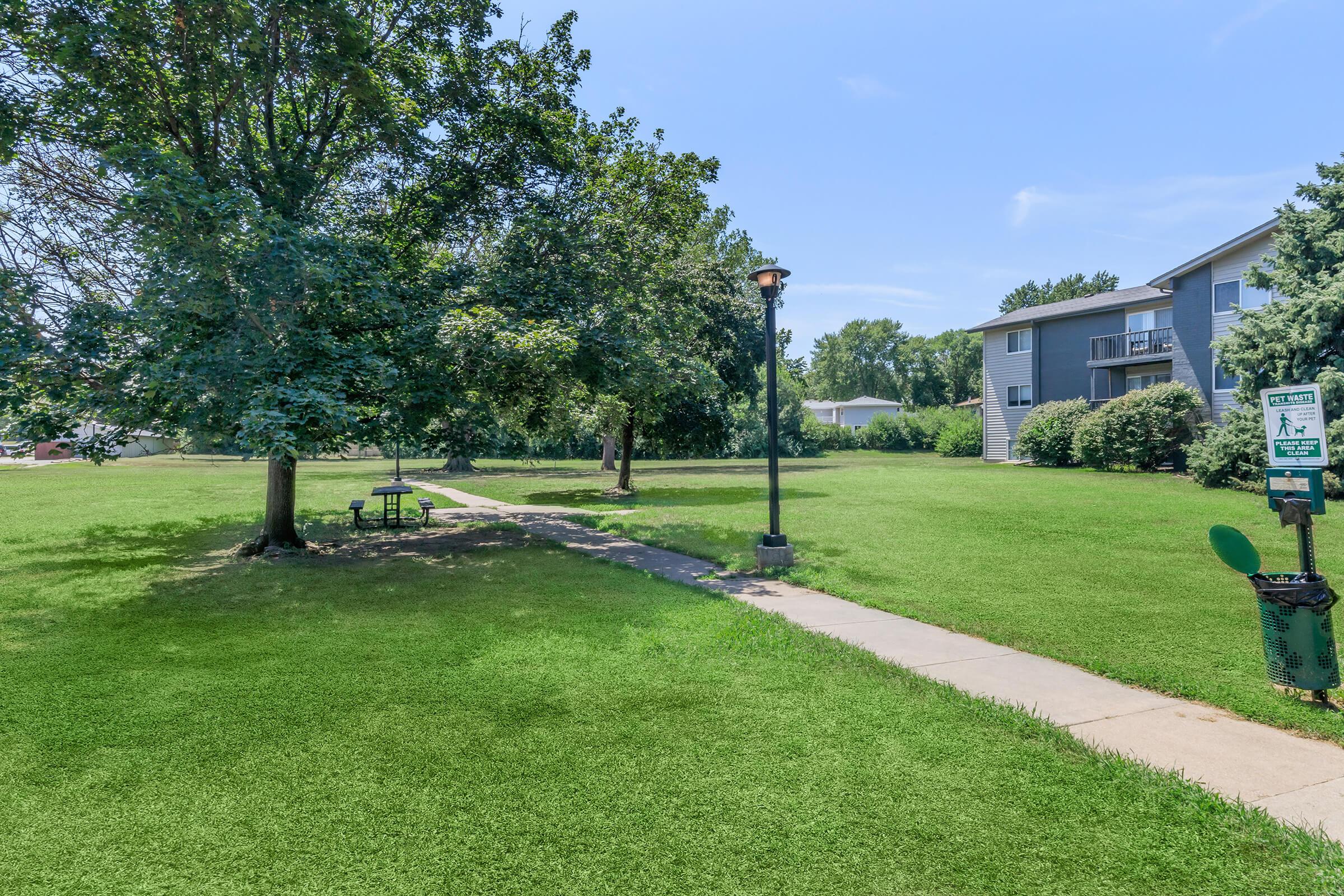 A grassy park area featuring two trees, a concrete path, and a lamppost. There is a picnic table on the grass, and an informational sign is visible nearby. In the background, an apartment building can be seen. The sky is blue with a few clouds.