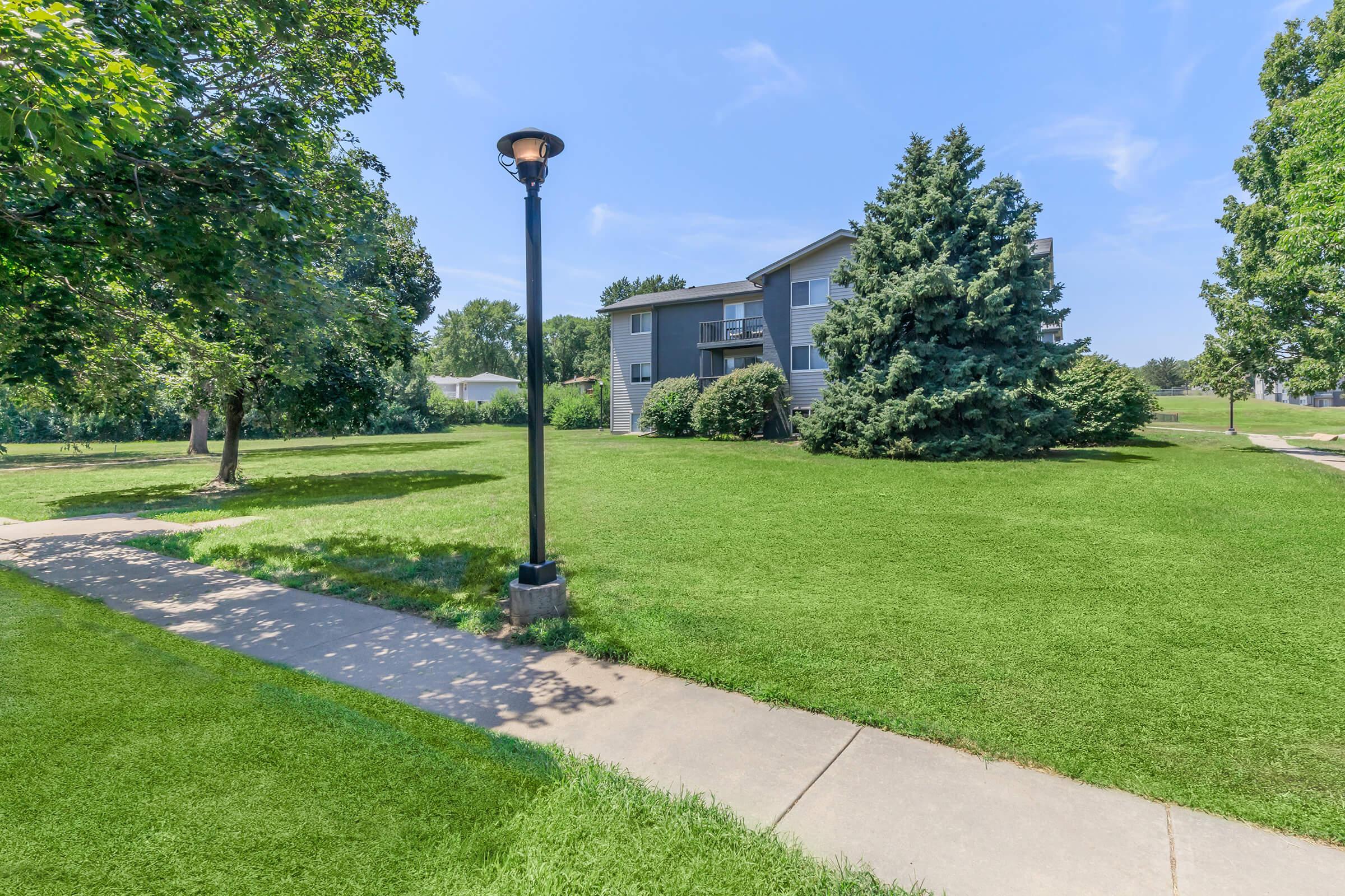 A well-maintained grassy area with a paved walkway leads to a multi-story residential building. The scene is framed by lush trees, and a lamppost adds to the inviting atmosphere under a clear blue sky.