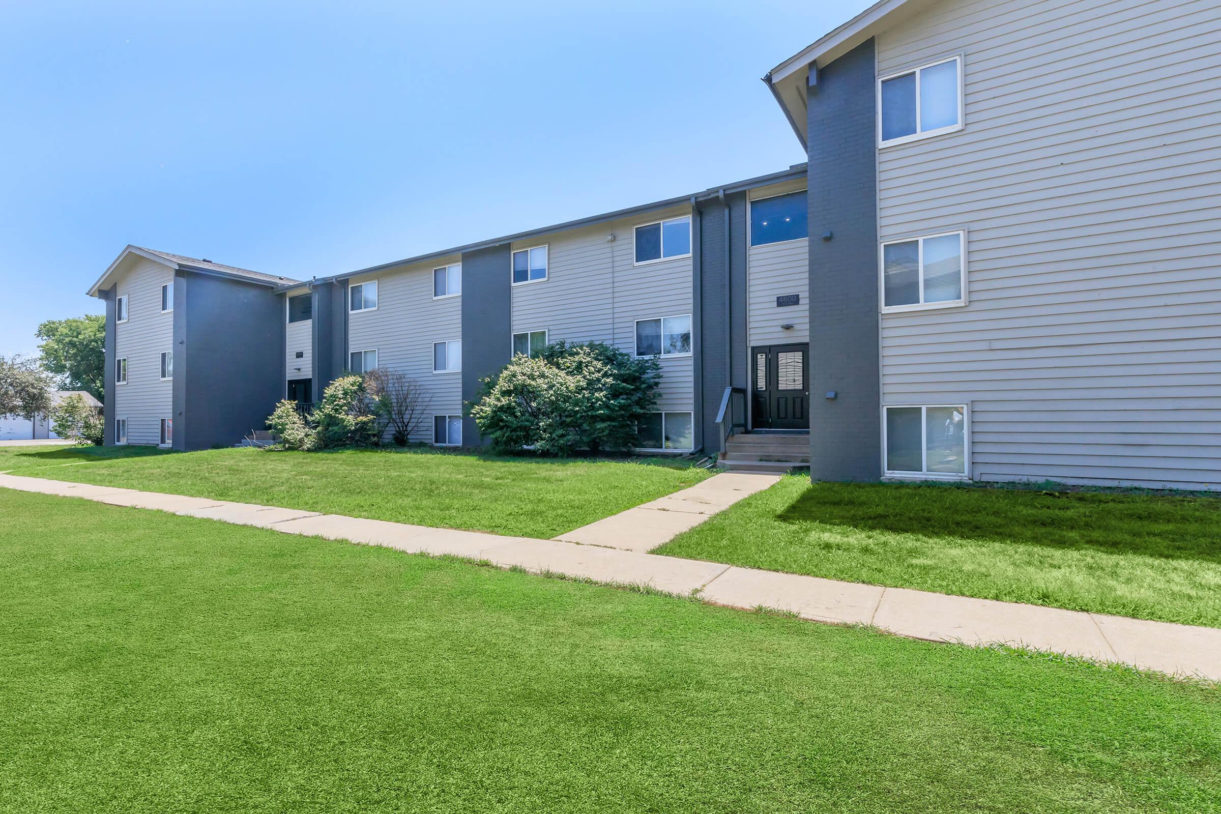 A modern, multi-unit residential building with gray siding, featuring multiple windows and a front walkway leading to the entrance. The lawn is well-maintained with lush green grass, and there are trees and shrubs planted around the exterior, under a clear blue sky.