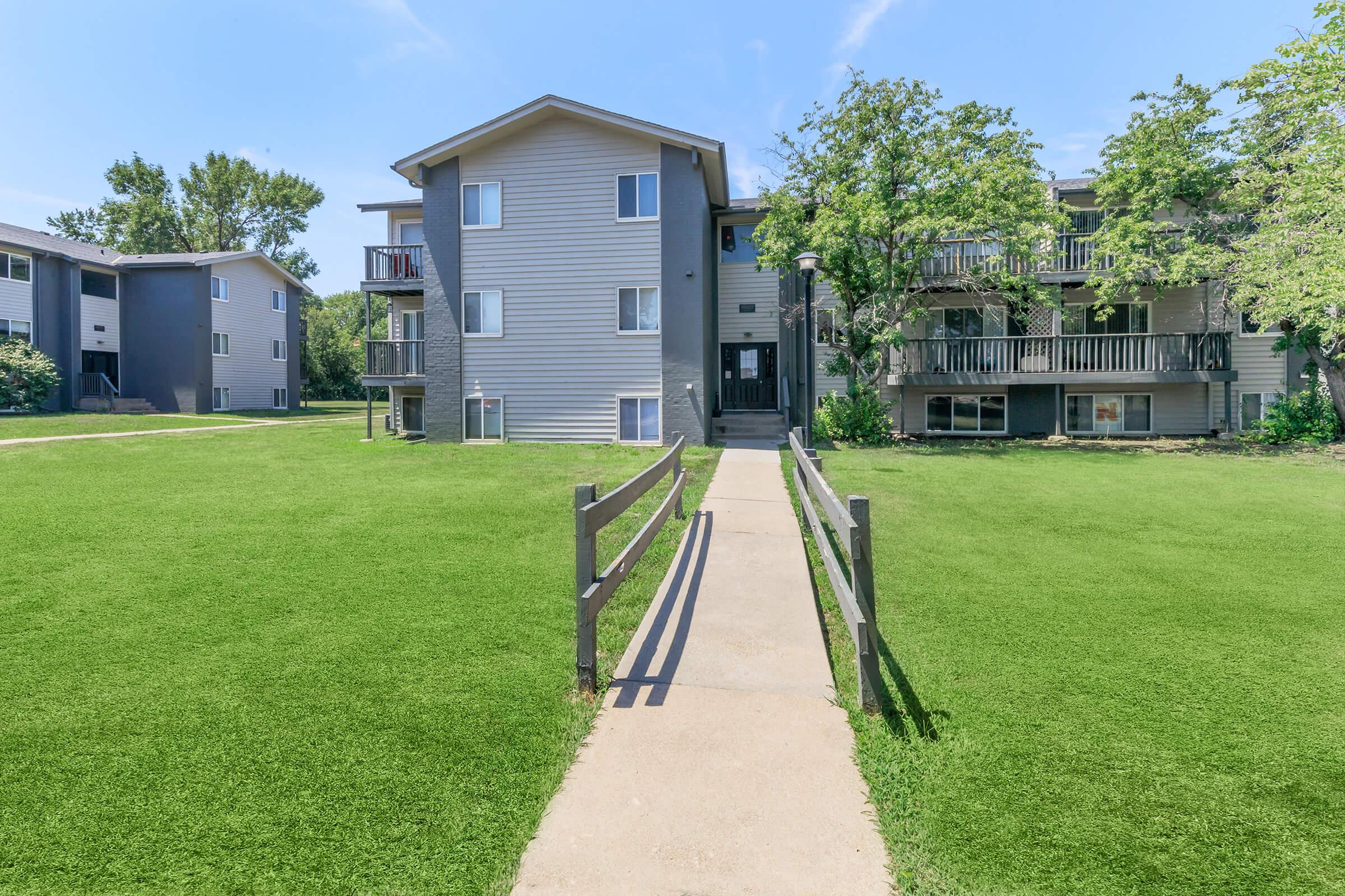 A view of a residential apartment complex with two buildings surrounded by green grass. A concrete pathway leads to the entrance, flanked by wooden fences and trees. The sky is clear and blue, creating a welcoming atmosphere.