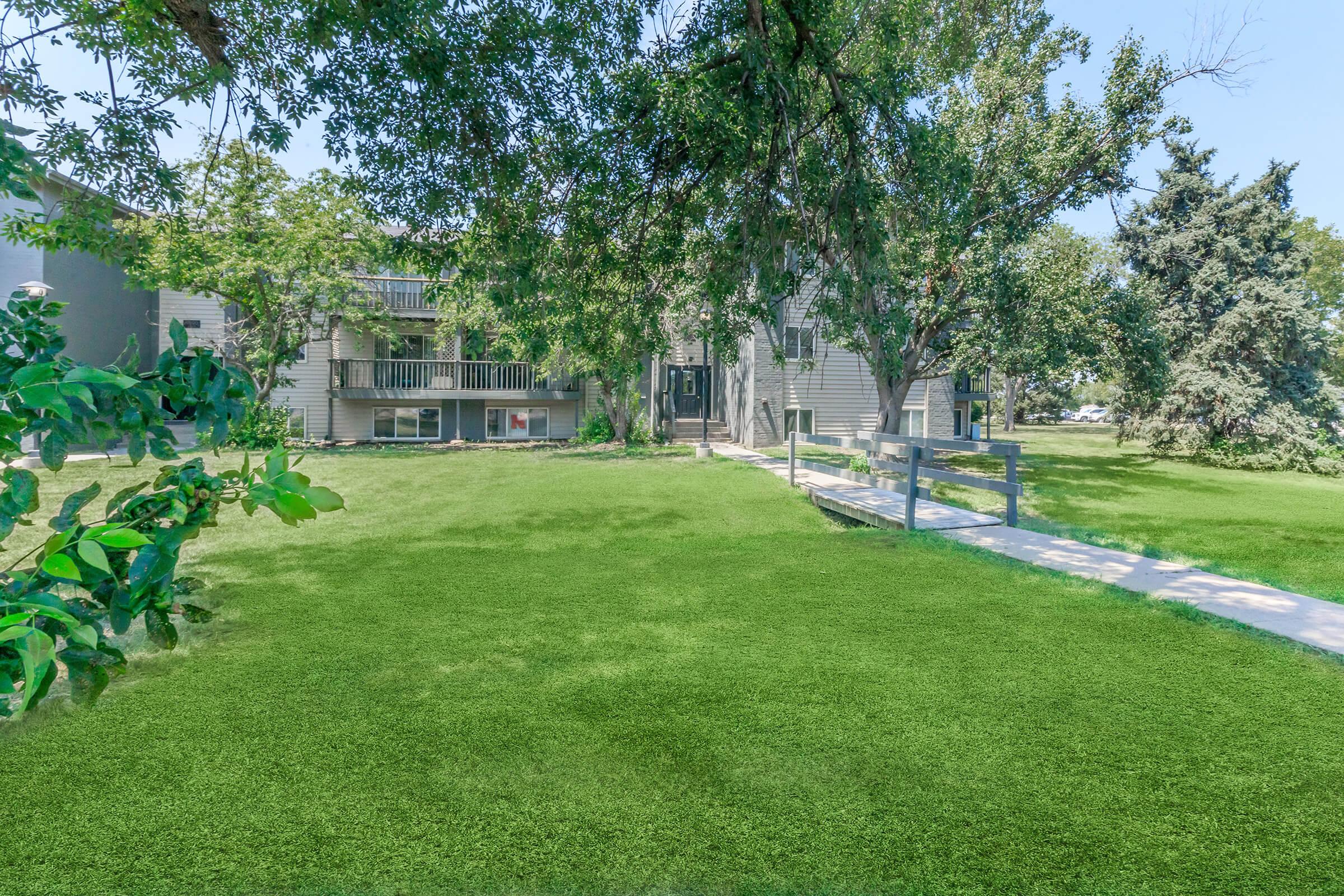 A well-maintained grassy area in front of a multi-story apartment building. The building features balconies, surrounded by trees and greenery, with a clear pathway leading to the entrance. The scene is bright and sunny, suggesting a pleasant outdoor environment.
